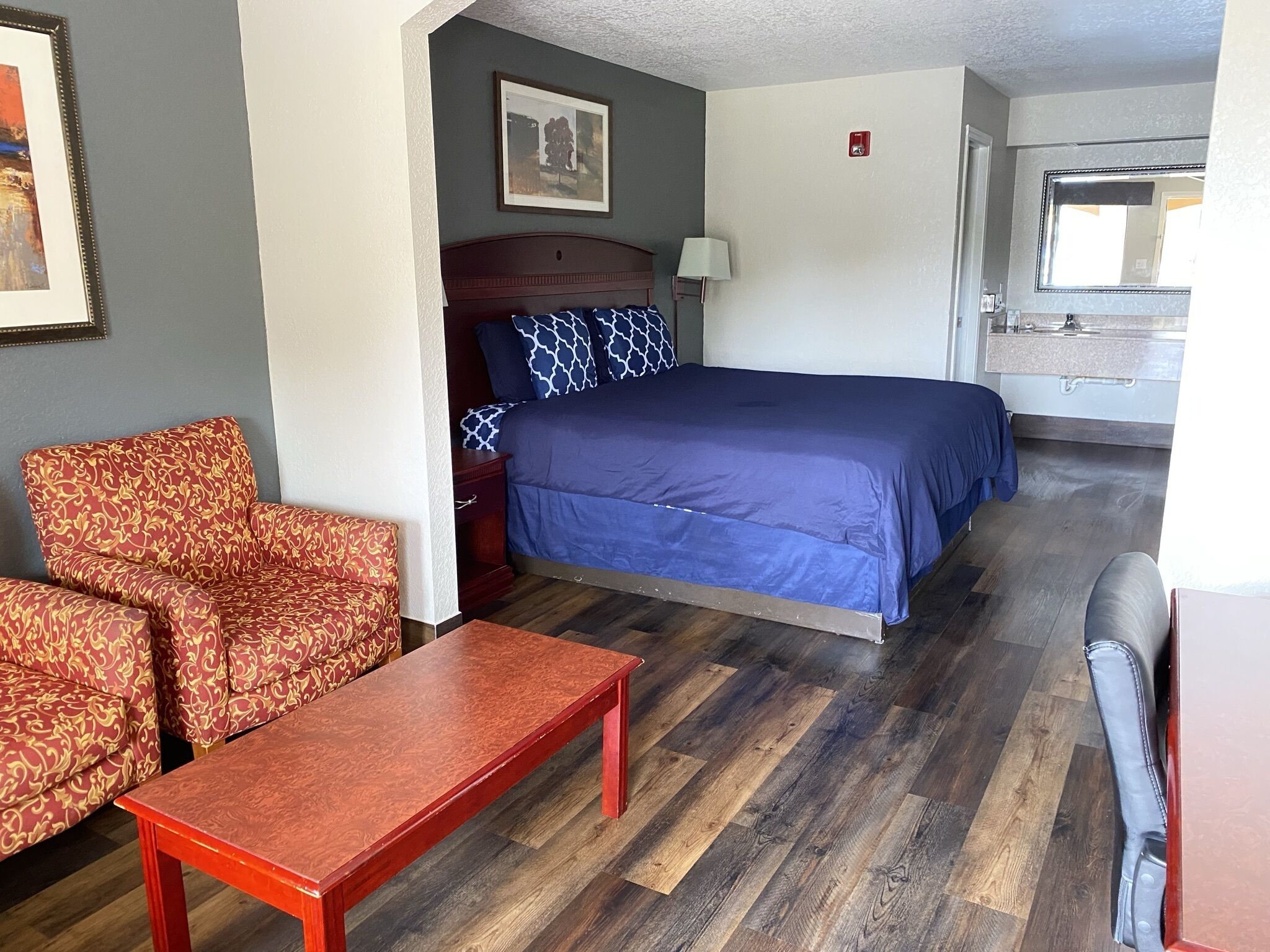 A hotel room with a bed, two patterned armchairs, a wooden table, and a view of the bathroom.