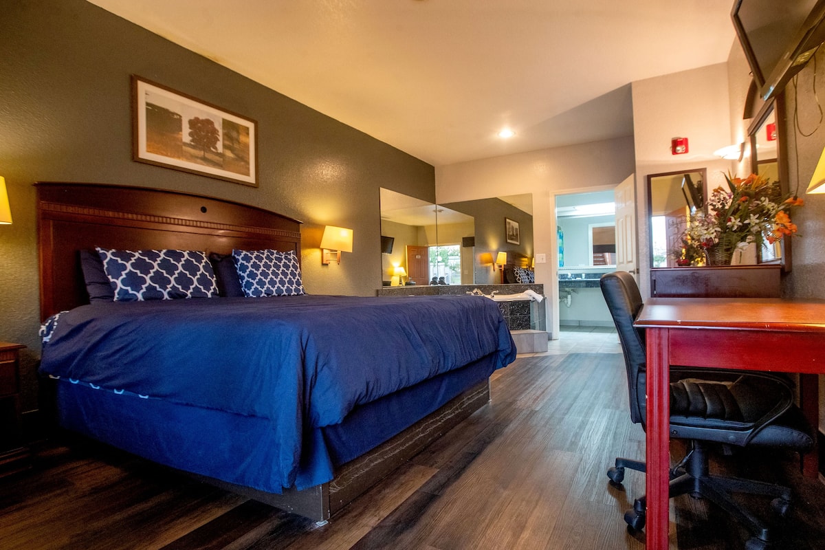 A hotel room with a large bed, a desk with a chair, a mirror, and a picture on the wall.