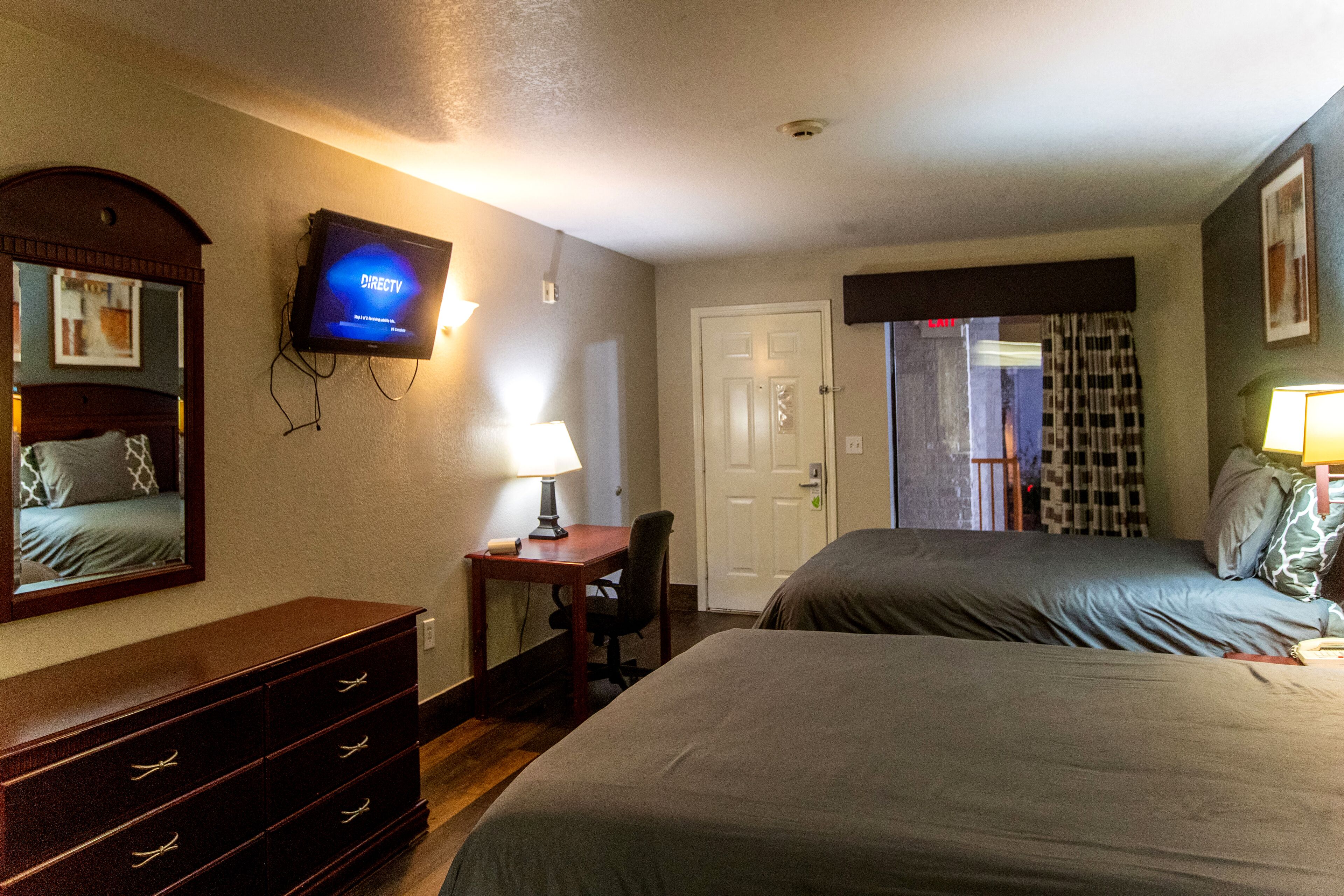 A hotel room with two beds, a desk with a lamp, a television mounted on the wall, and a door leading to another room.