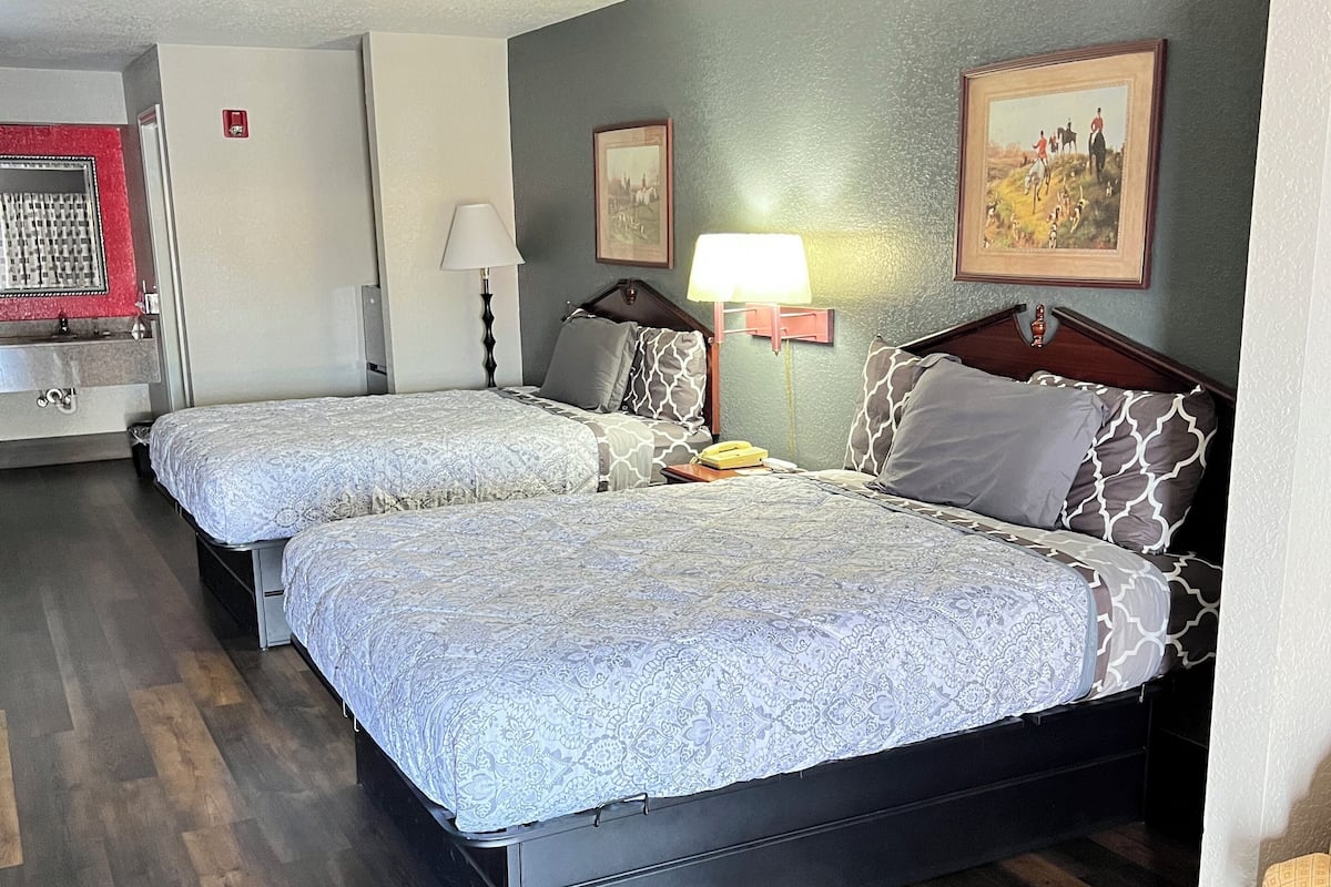 A hotel room with two beds, a wooden floor, a desk, a chair, a lamp, and a painting on the wall.