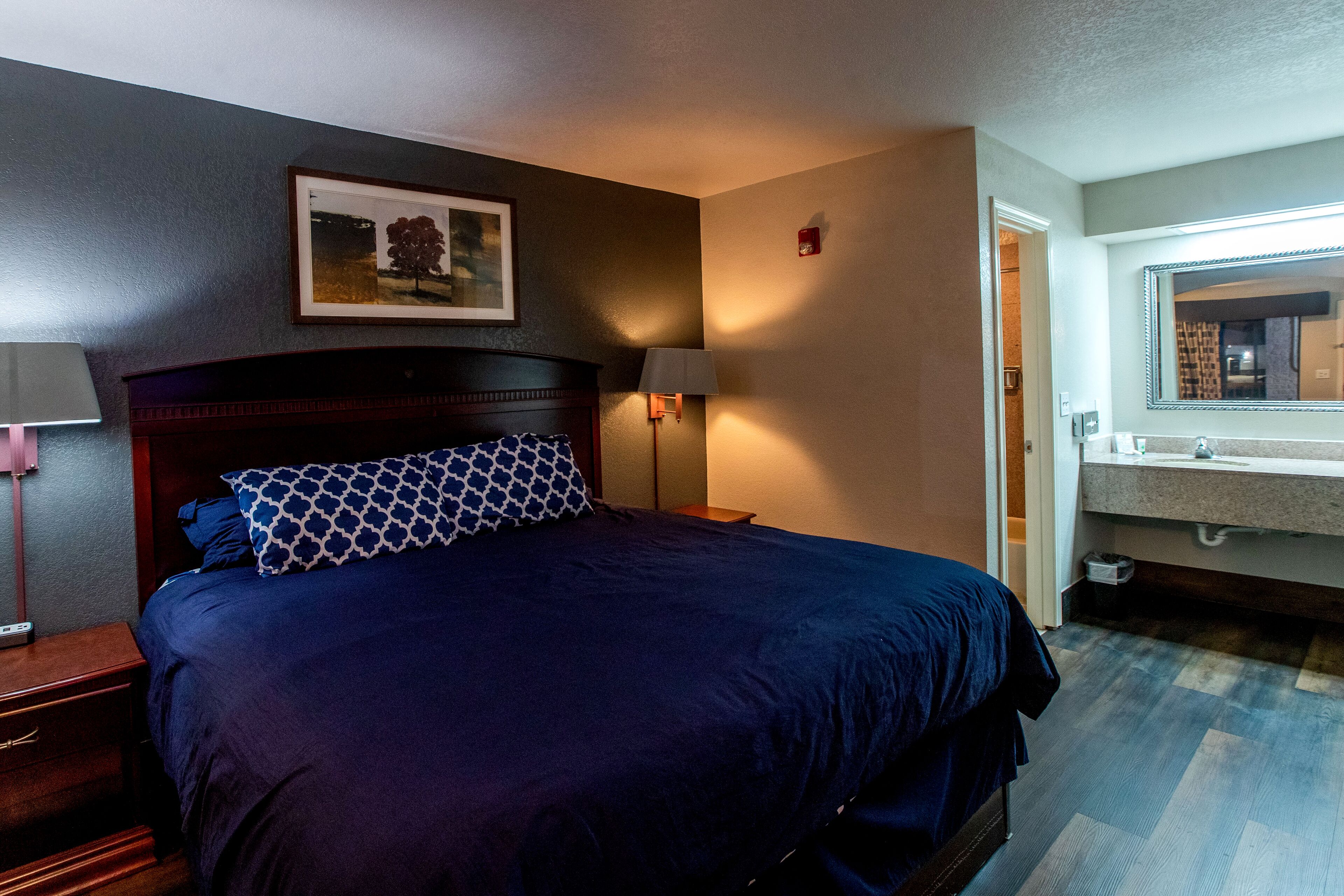 Ein Hotelzimmer mit einem großen Bett, einem Nachttisch, einer Lampe, einem gerahmten Bild an der Wand und einem Badezimmer mit Spiegel und Waschbecken.