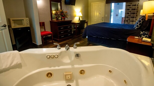 A hotel room with a large bathtub, a desk with a lamp, a television, and a bed with a blue blanket. - Magnolia Inn And Suites (Magnolia)