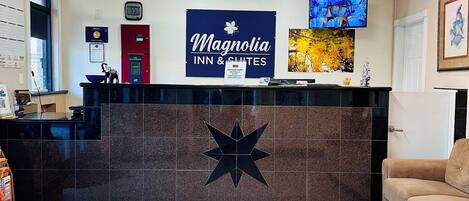 Zona de recepción de un hotel con un cartel grande que dice Magnolia Inn & Suites, un reloj digital, un televisor montado en la pared y piso de baldosas.