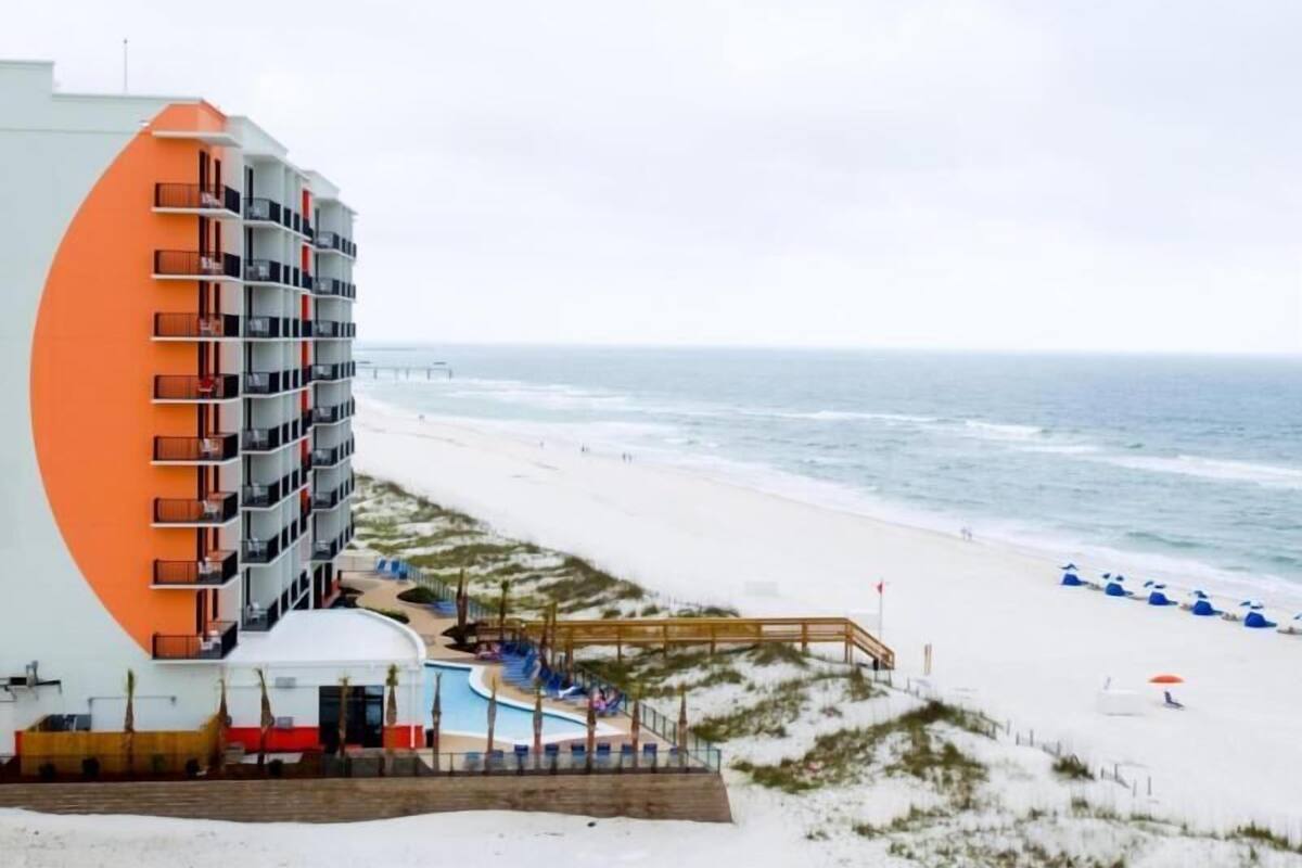 A beachfront hotel with a large building featuring balconies, a pool area, and a beach with umbrellas.