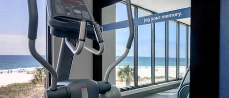 A gym with a treadmill and exercise equipment, overlooking a beach with palm trees and a clear sky.
