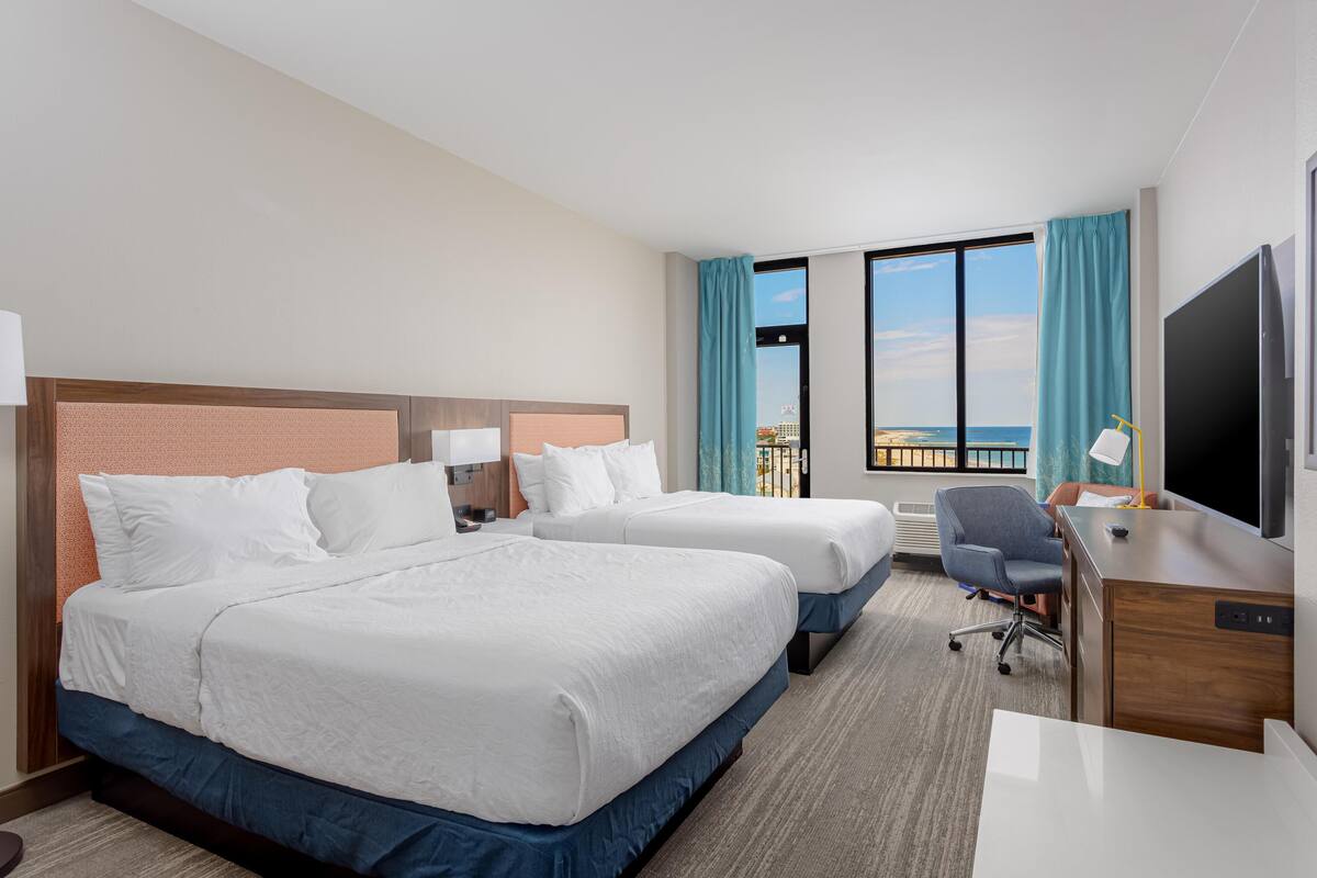 A hotel room with two beds, a desk, a chair, a TV, and a large window with a view of the sea.