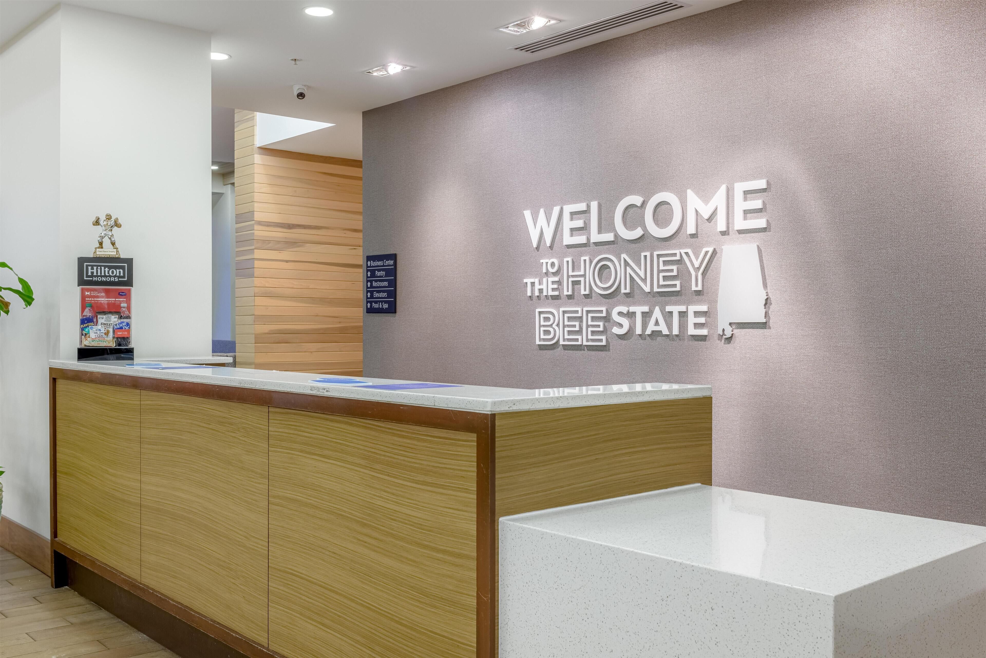 A reception desk with a wooden finish and a white countertop. A sign on the wall reads "WELCOME TO THE HONEY BEE STATE."