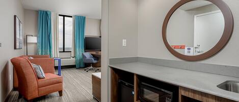 A hotel room with a sofa, a desk, a chair, a large mirror, a built-in oven, and a sink.