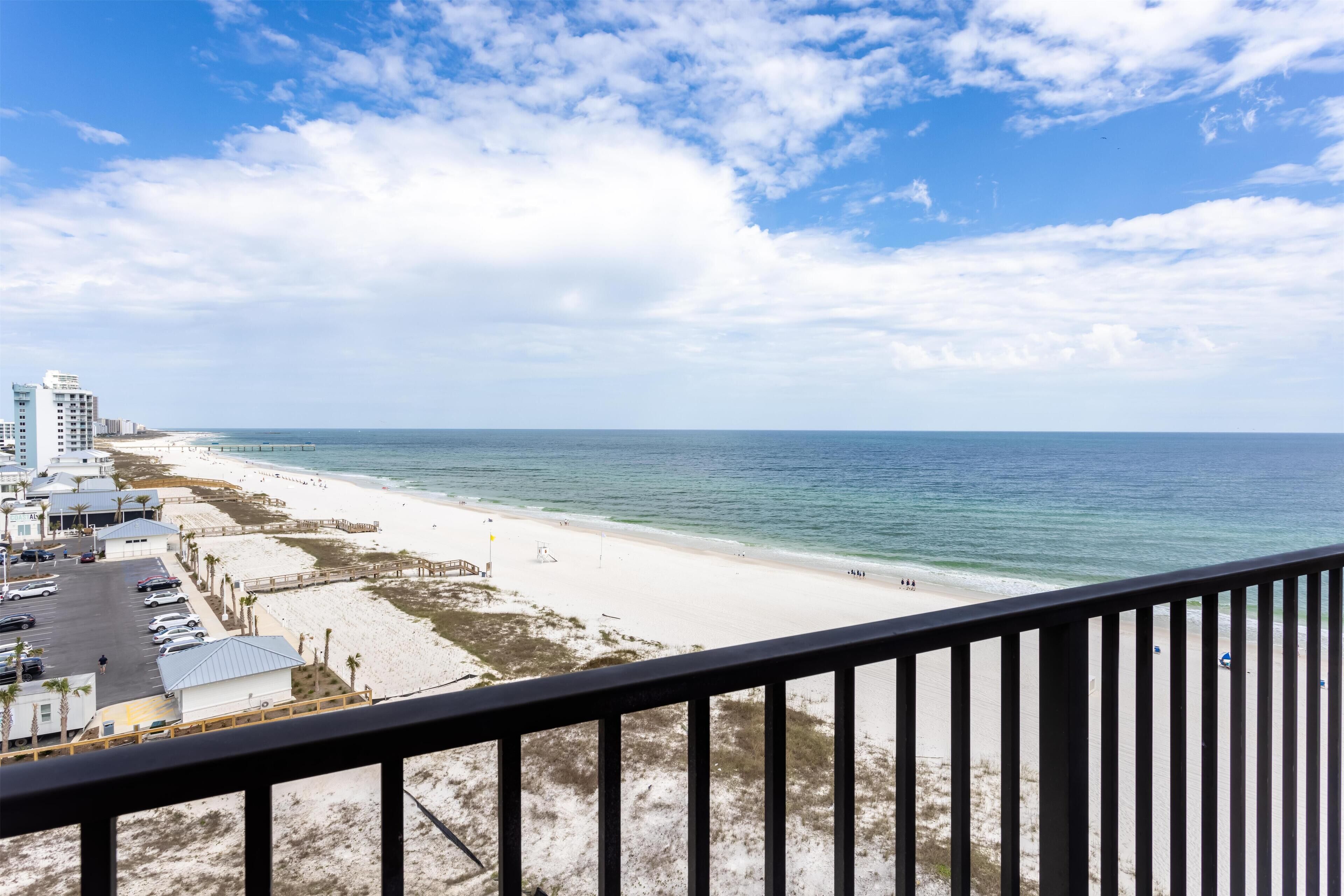 A beach view from a balcony with a railing, sandy beach, ocean, and buildings in the background.