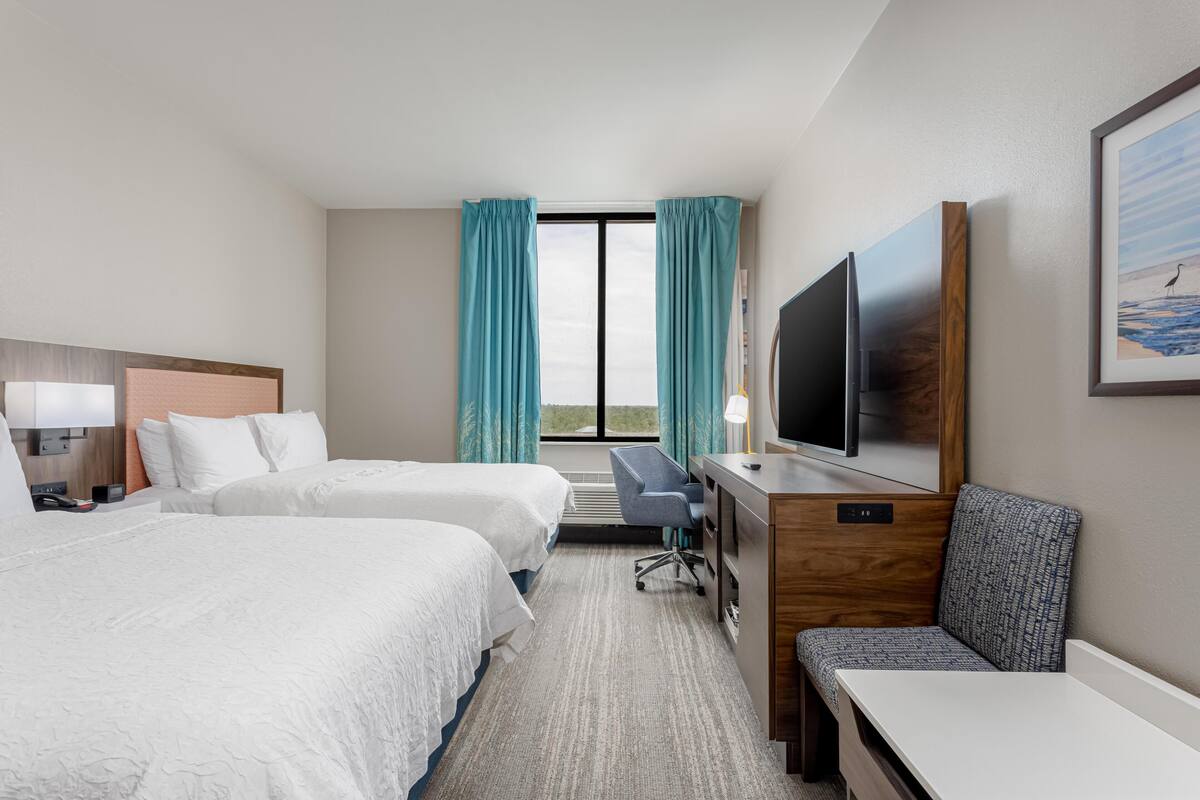 A hotel room with two beds, a desk, a chair, a TV, and a large window with curtains.