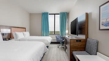 A hotel room with two beds, a desk, a chair, a TV, and a large window with curtains.