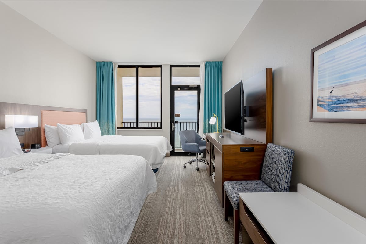 A hotel room with two beds, a desk, a chair, a TV, and a large window with curtains.