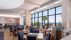 Uma zona de refeições moderna com mesas e cadeiras, grandes janelas com vista para o mar, e um espaço de estar informal com sofás e uma pequena mesa.