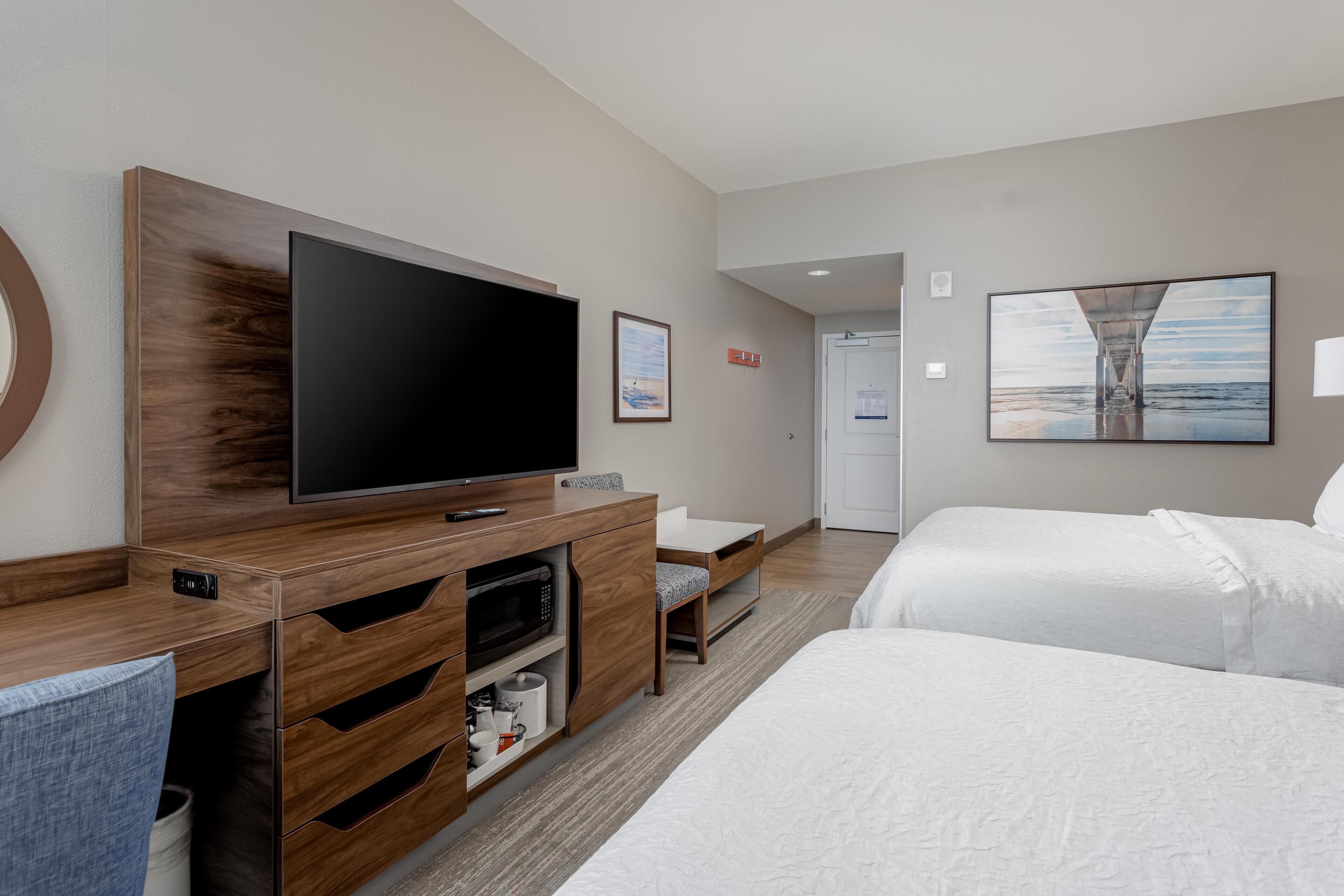 Una habitación de hotel moderna con un televisor de pantalla plana grande, un mueble de madera para entretenimiento, una cama, un escritorio y un cuadro en la pared.