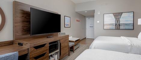 A modern hotel room with a large flat-screen TV, a wooden entertainment unit, a bed, a desk, and a framed picture on the wall.