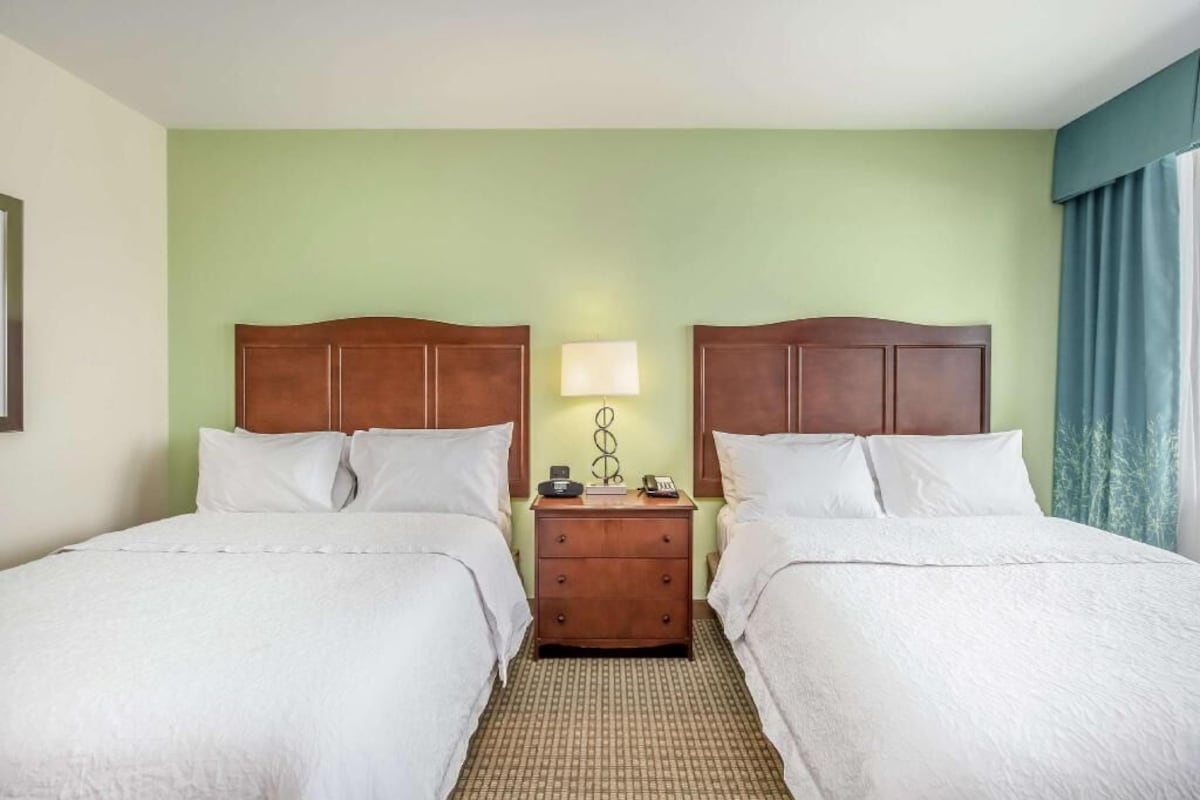 A hotel room with two beds, a nightstand with a lamp, and a window with curtains.