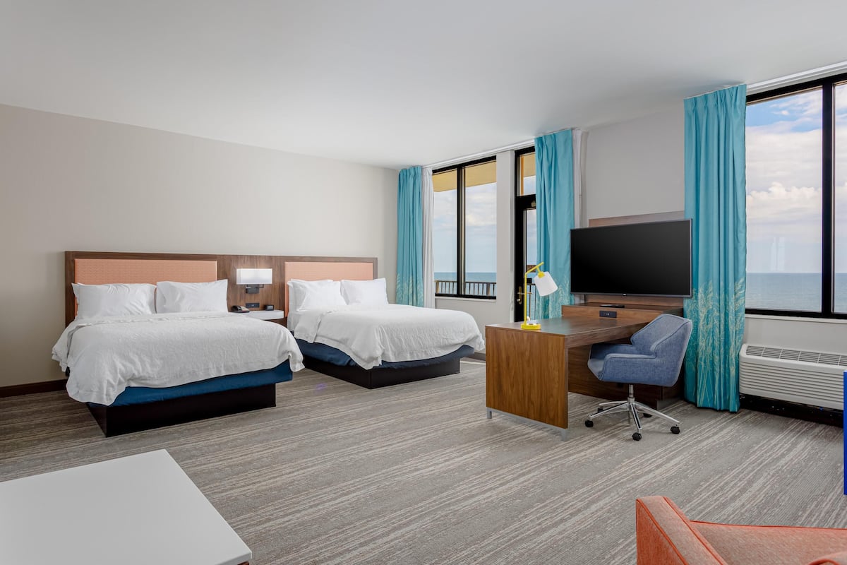 A hotel room with two beds, a desk, a chair, a TV, and a large window with curtains.