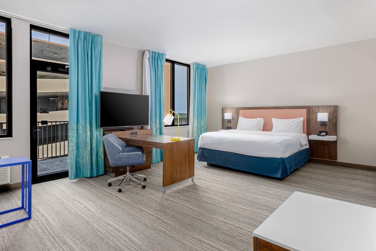 A hotel room with a bed, desk, chair, TV, and a balcony view.