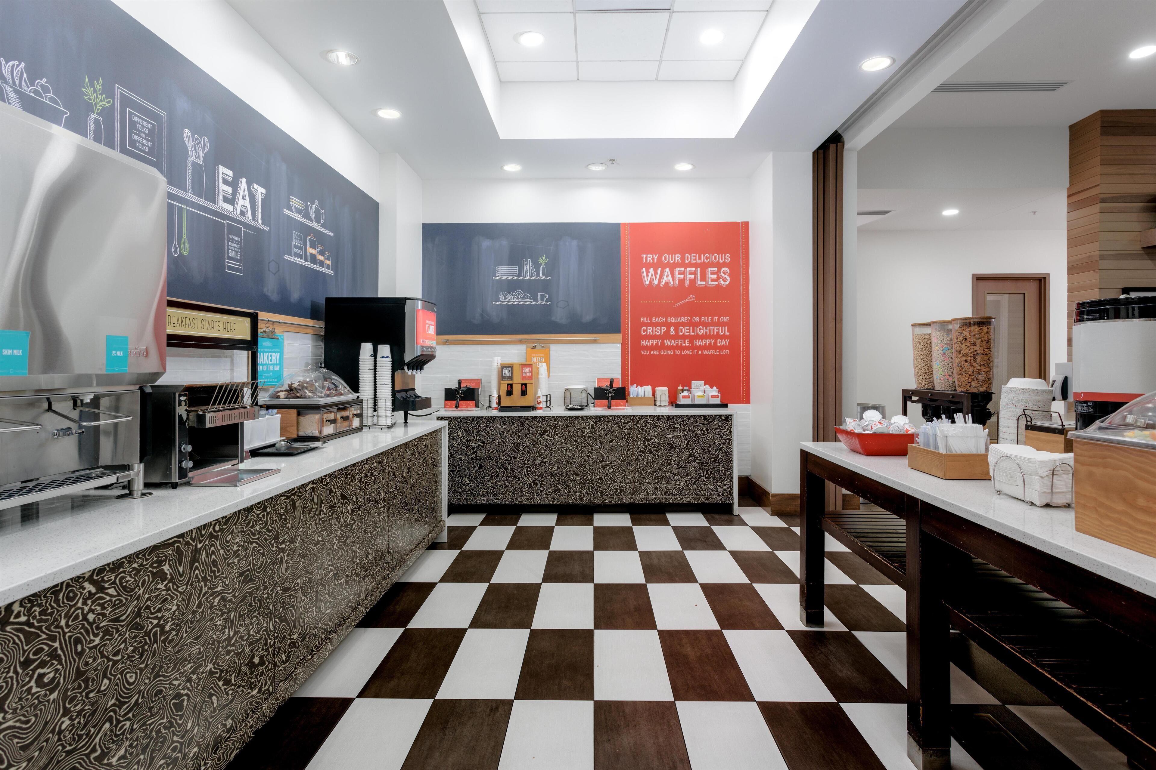A modern breakfast area with a checkered floor, a counter with various food items, and a wall with a 'Breakfast Starts Here' sign.