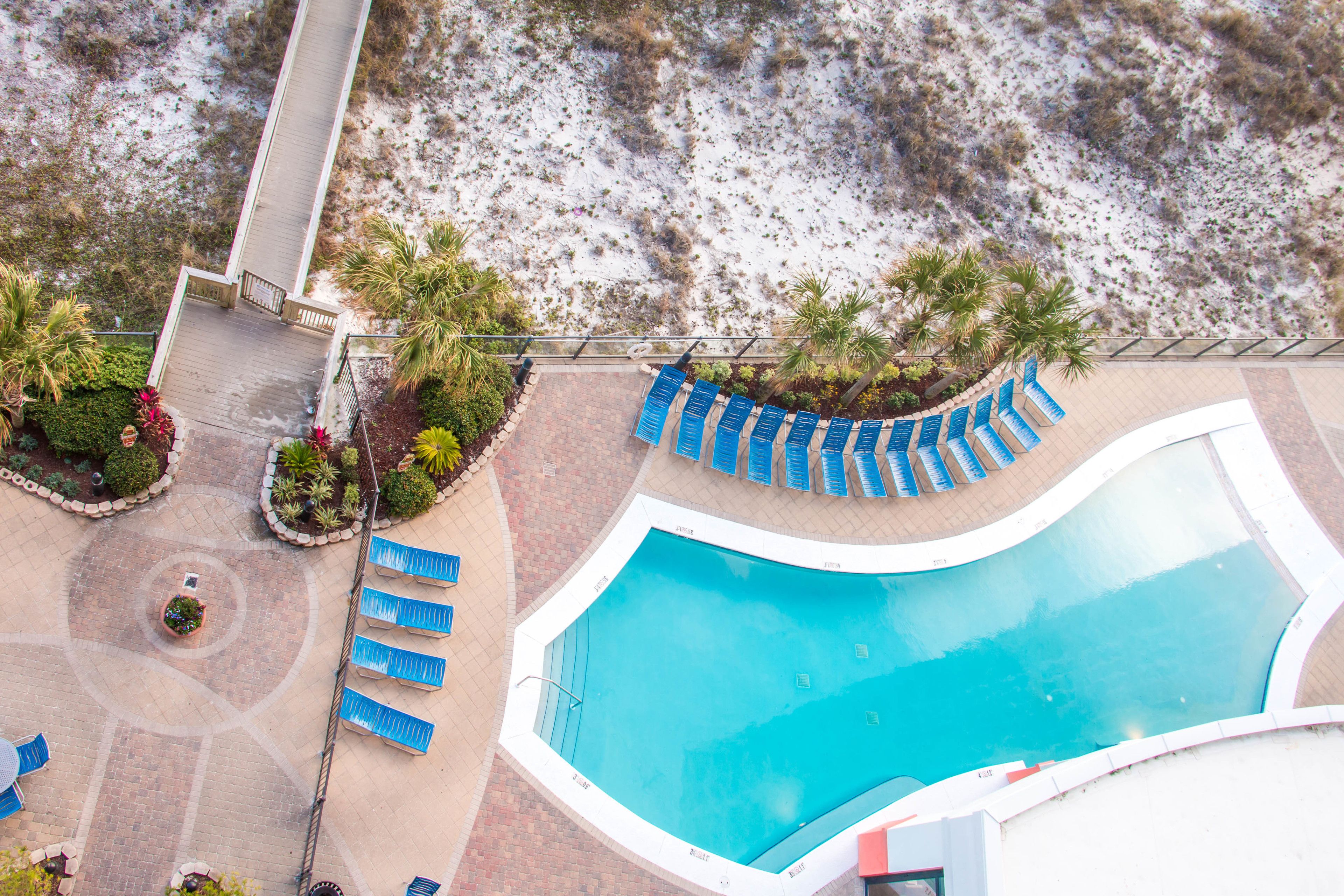 Vista aérea de un complejo turístico con piscina, sillas de descanso y palmeras.
