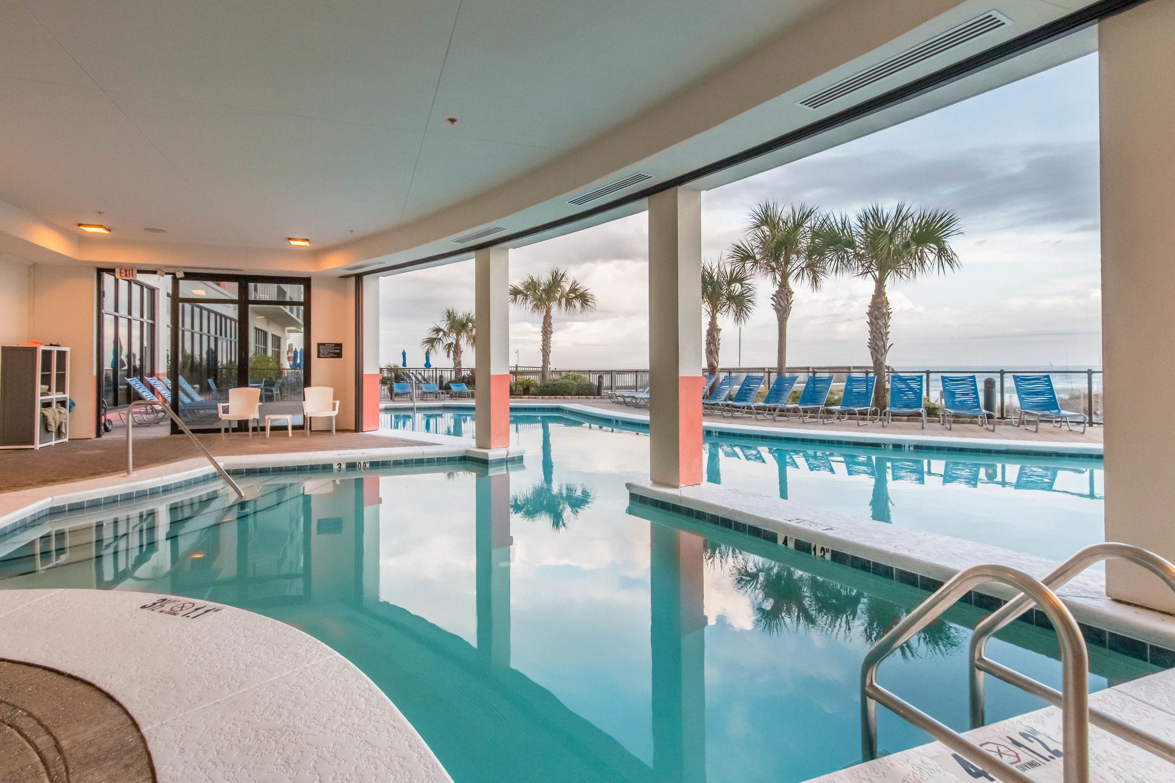 An indoor swimming pool with clear blue water, surrounded by lounge chairs and palm trees, and a view of the ocean through large windows.