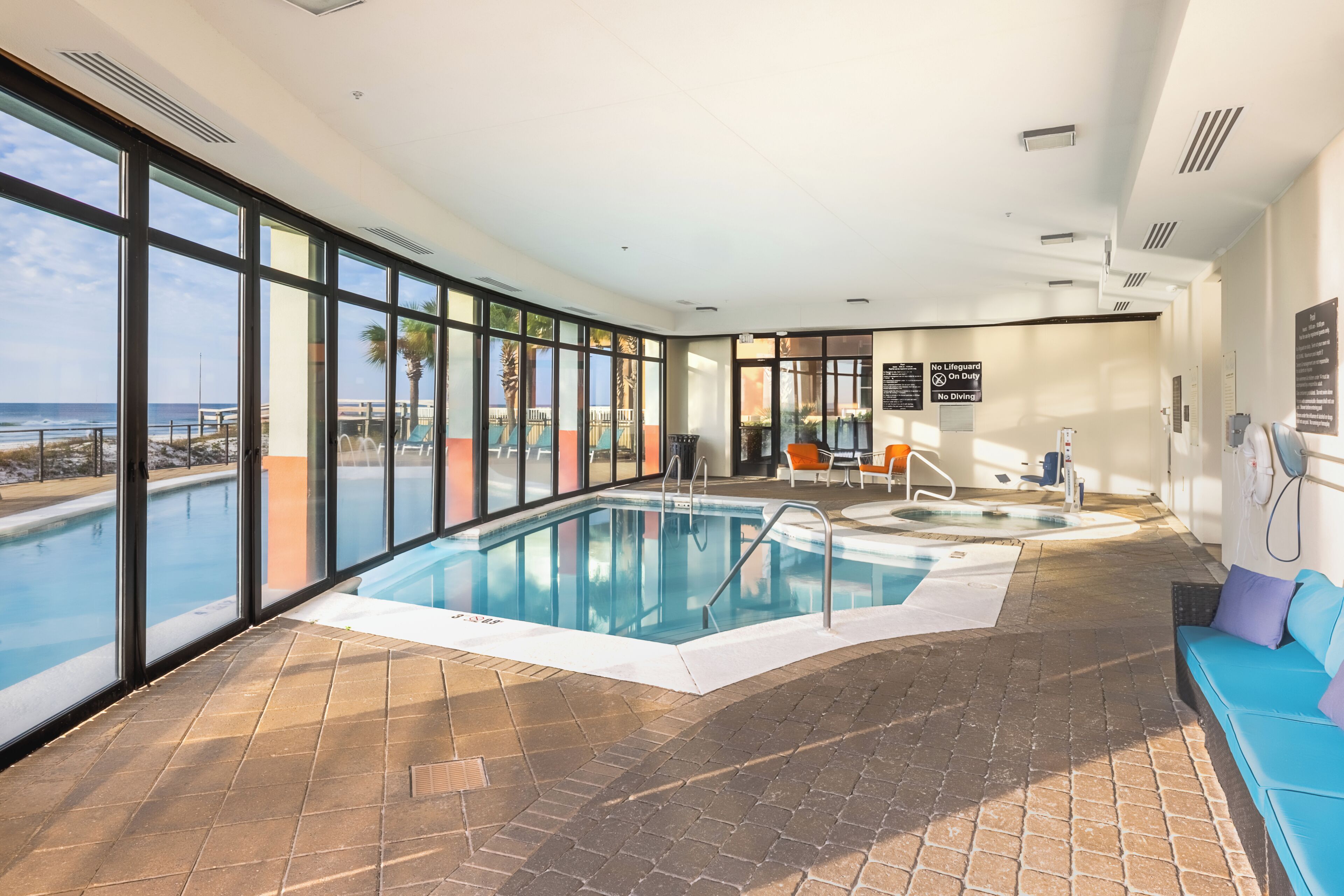 An indoor swimming pool with clear views of the ocean, a lounge area, and a no-lifeguard sign.