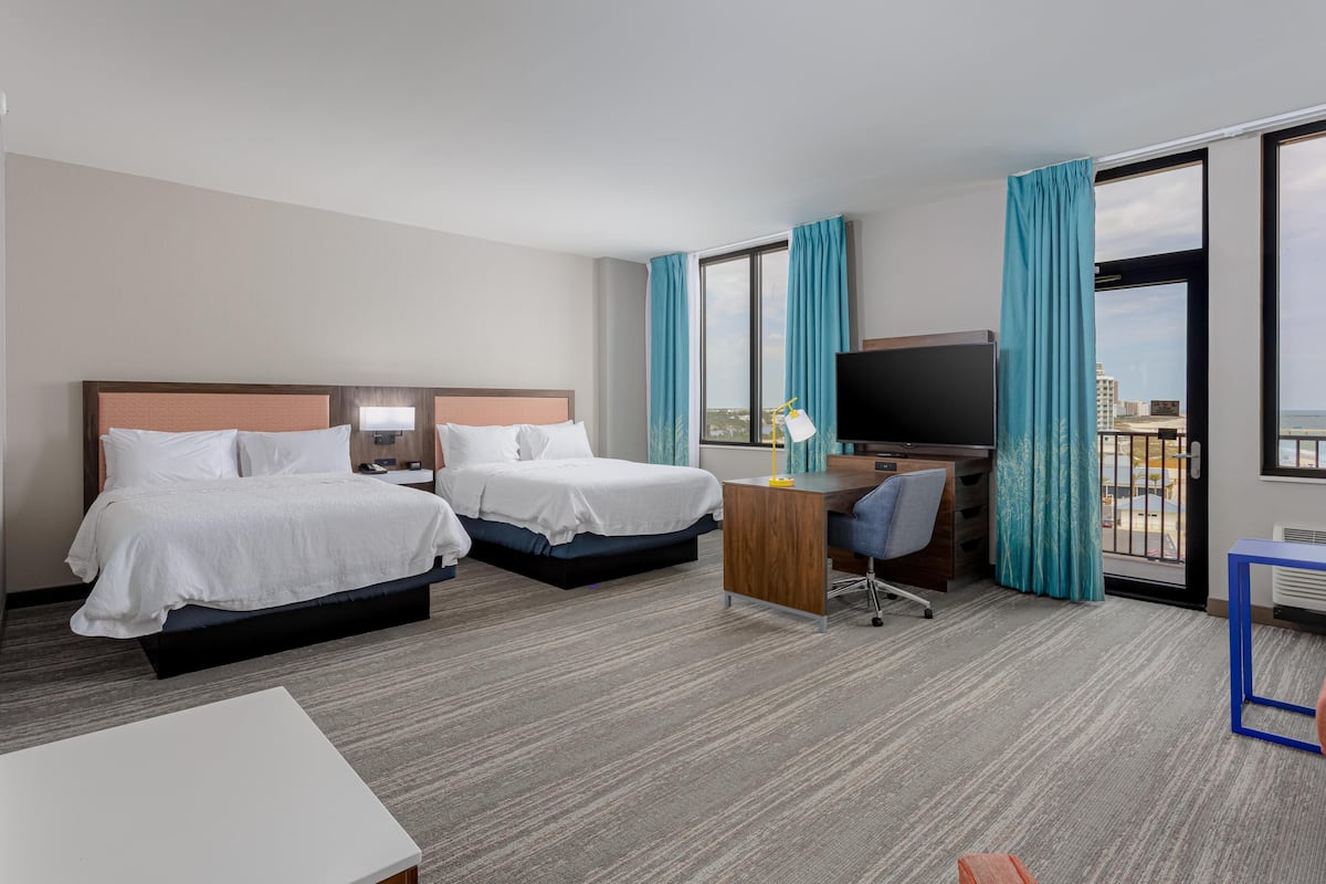 A hotel room with two beds, a desk, a TV, and large windows with curtains.