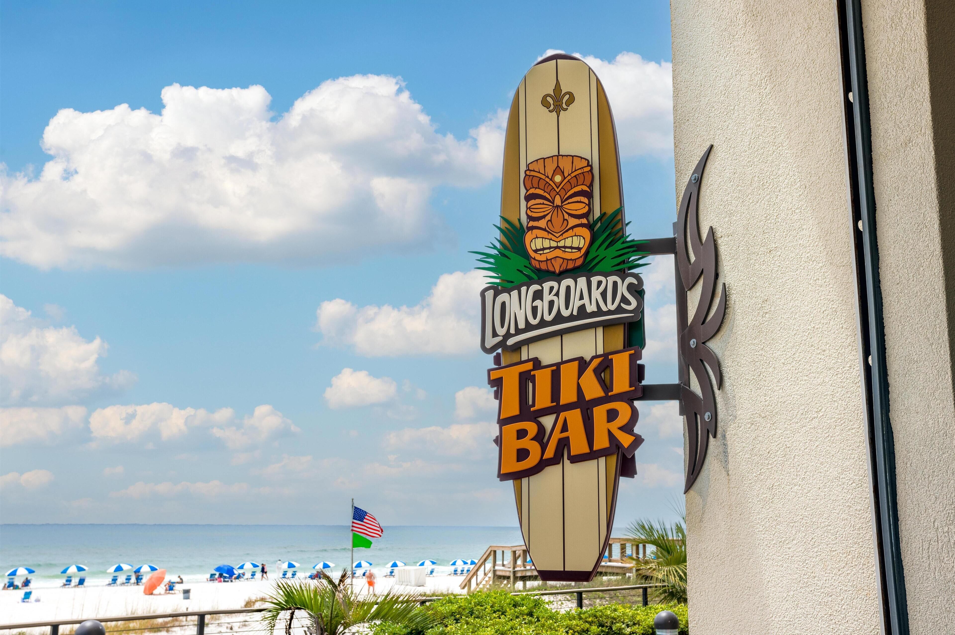 A surfboard-shaped sign for Longboards Tiki Bar with a tiki mask design, located near a beach with umbrellas and a flag.