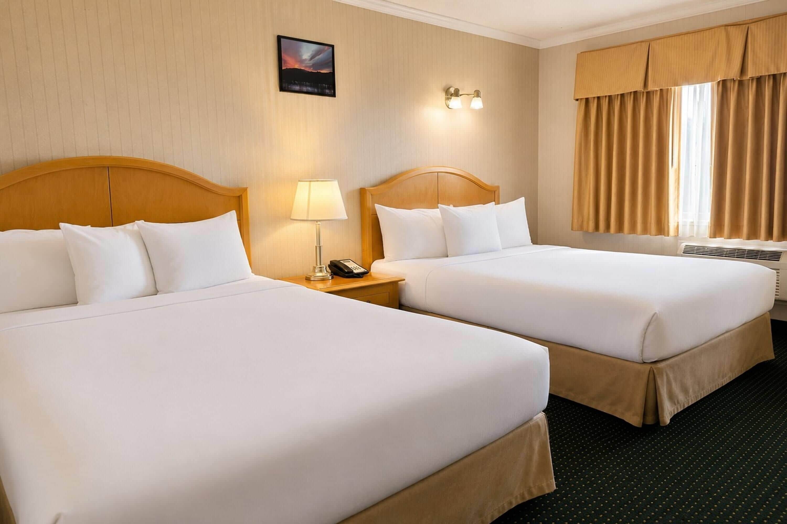 A hotel room with two beds, a nightstand with a lamp, a phone, and a wall-mounted light fixture.