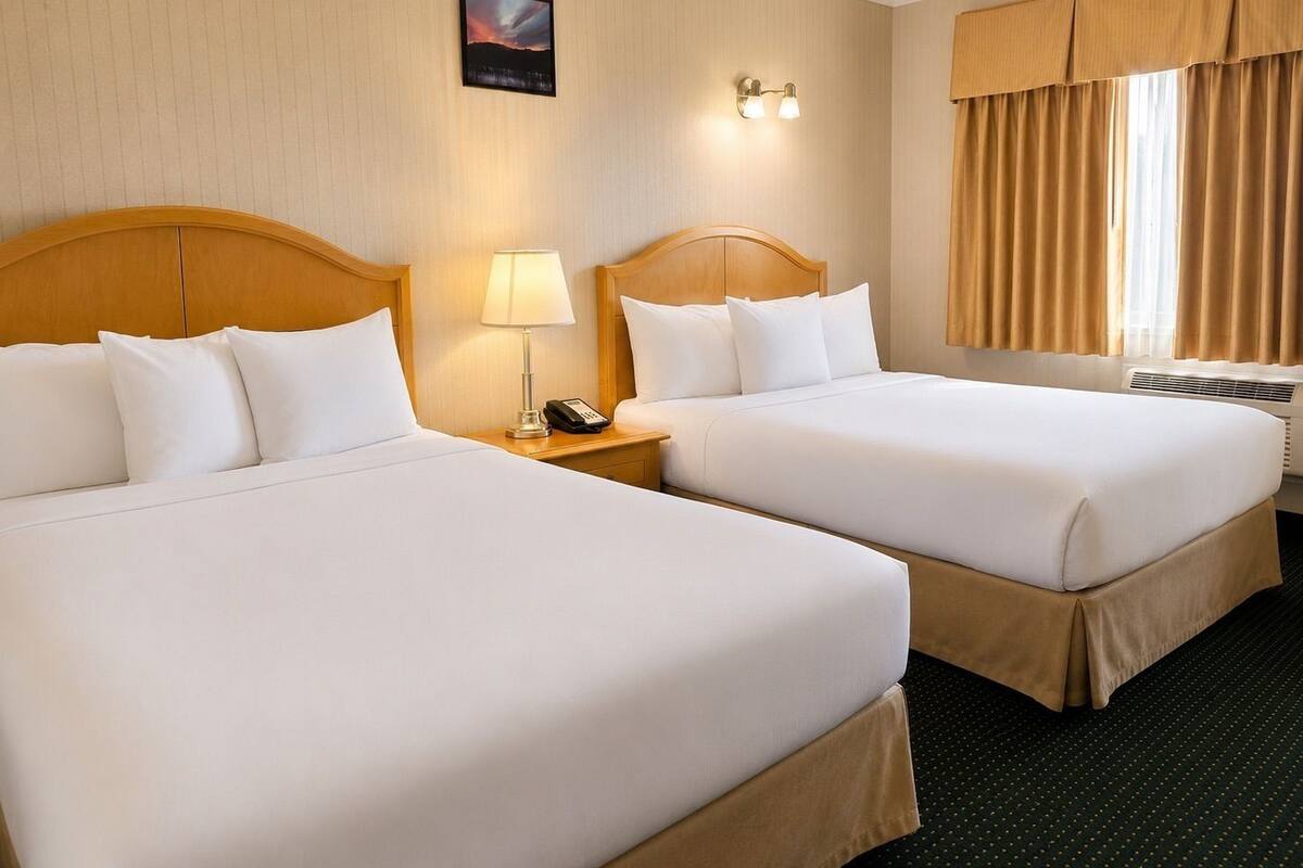 Habitación de hotel con dos camas, una mesita de noche con lámpara y teléfono, una luz en la pared y una ventana con cortinas.