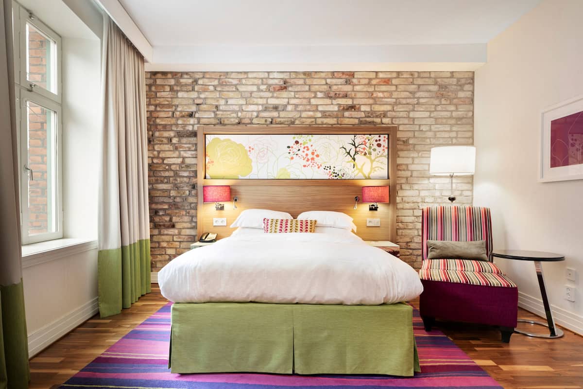 Hotellihuone, jossa on tiiliseinä, vihreällä kankaalla verhoiltu sängynpääty, raidallinen nojatuoli ja pieni pyöreä pöytä.