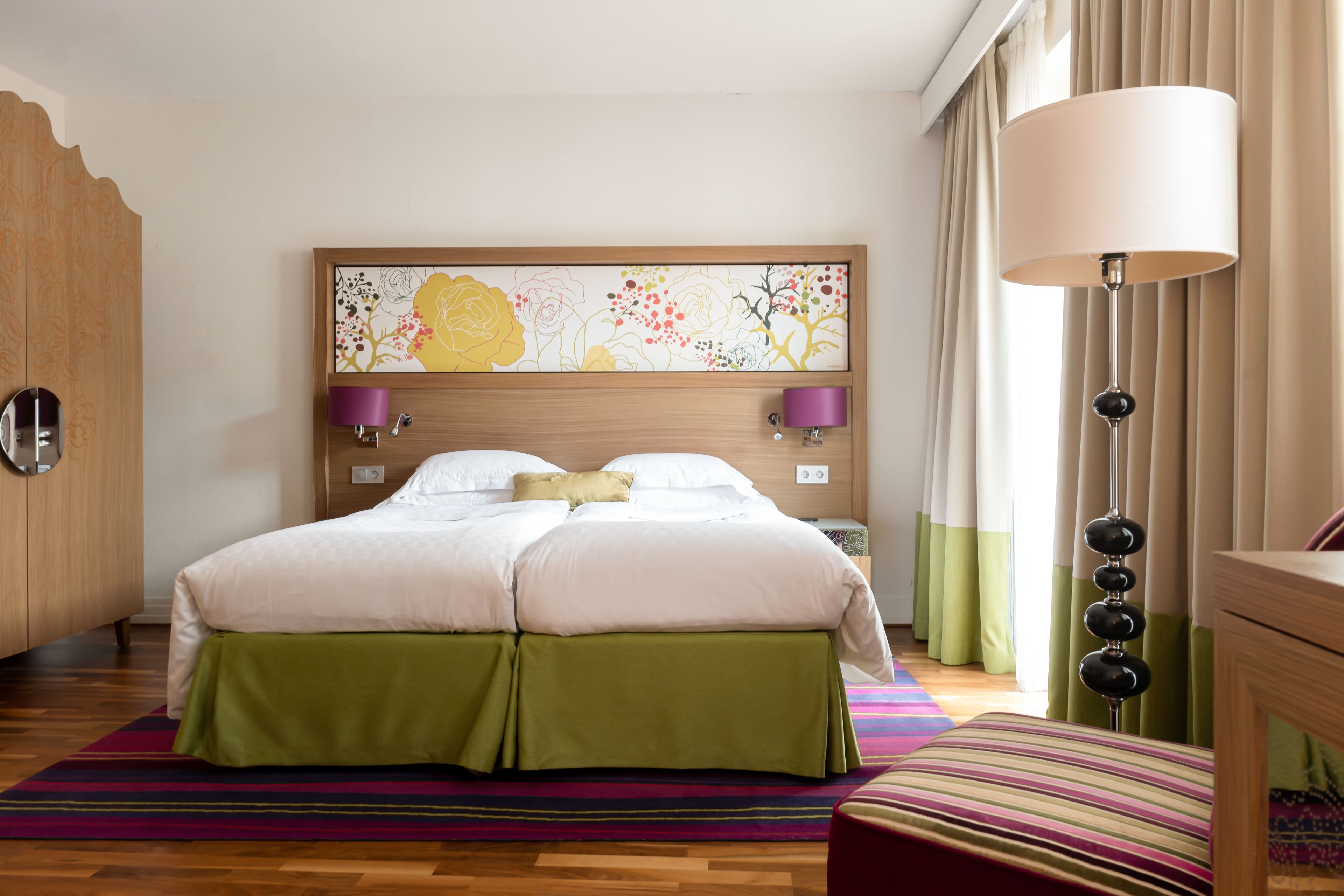 A hotel room with a bed, a headboard with a floral artwork, a bedside lamp, and a green skirted bed.