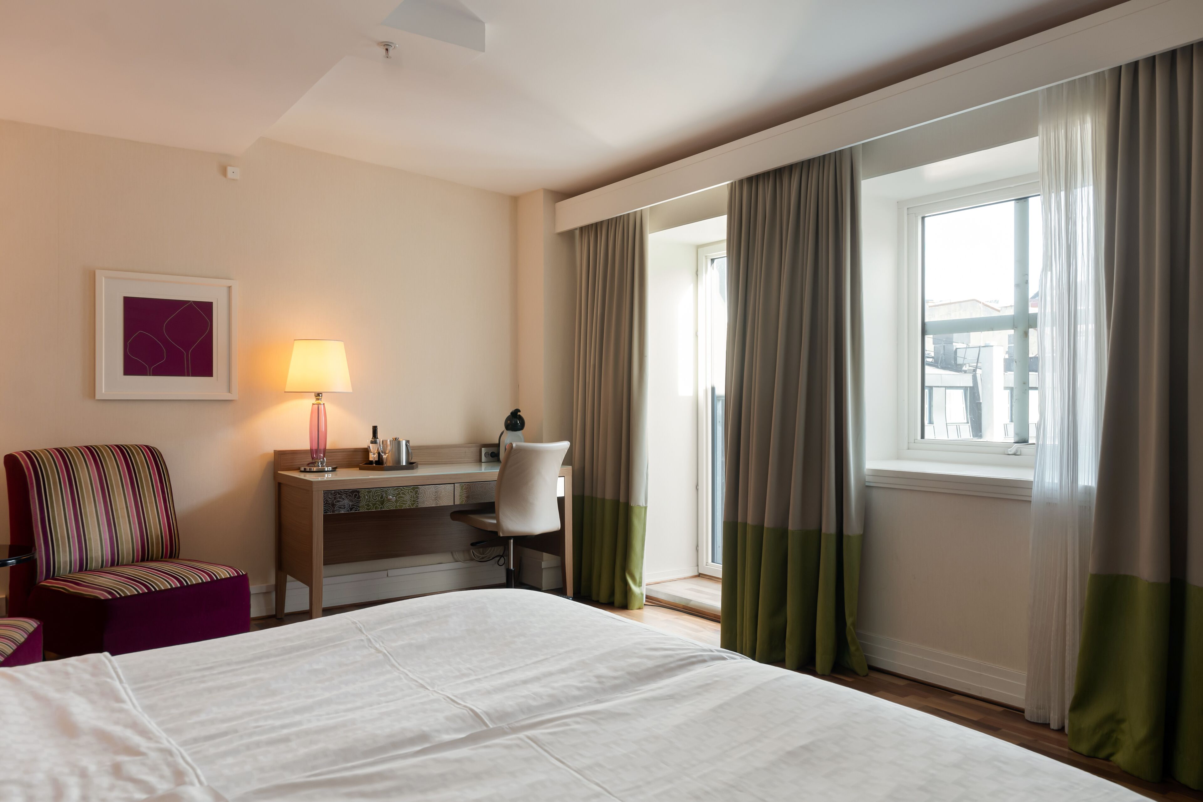 A hotel room with a bed, a desk with a chair, a lamp, and a window with curtains.