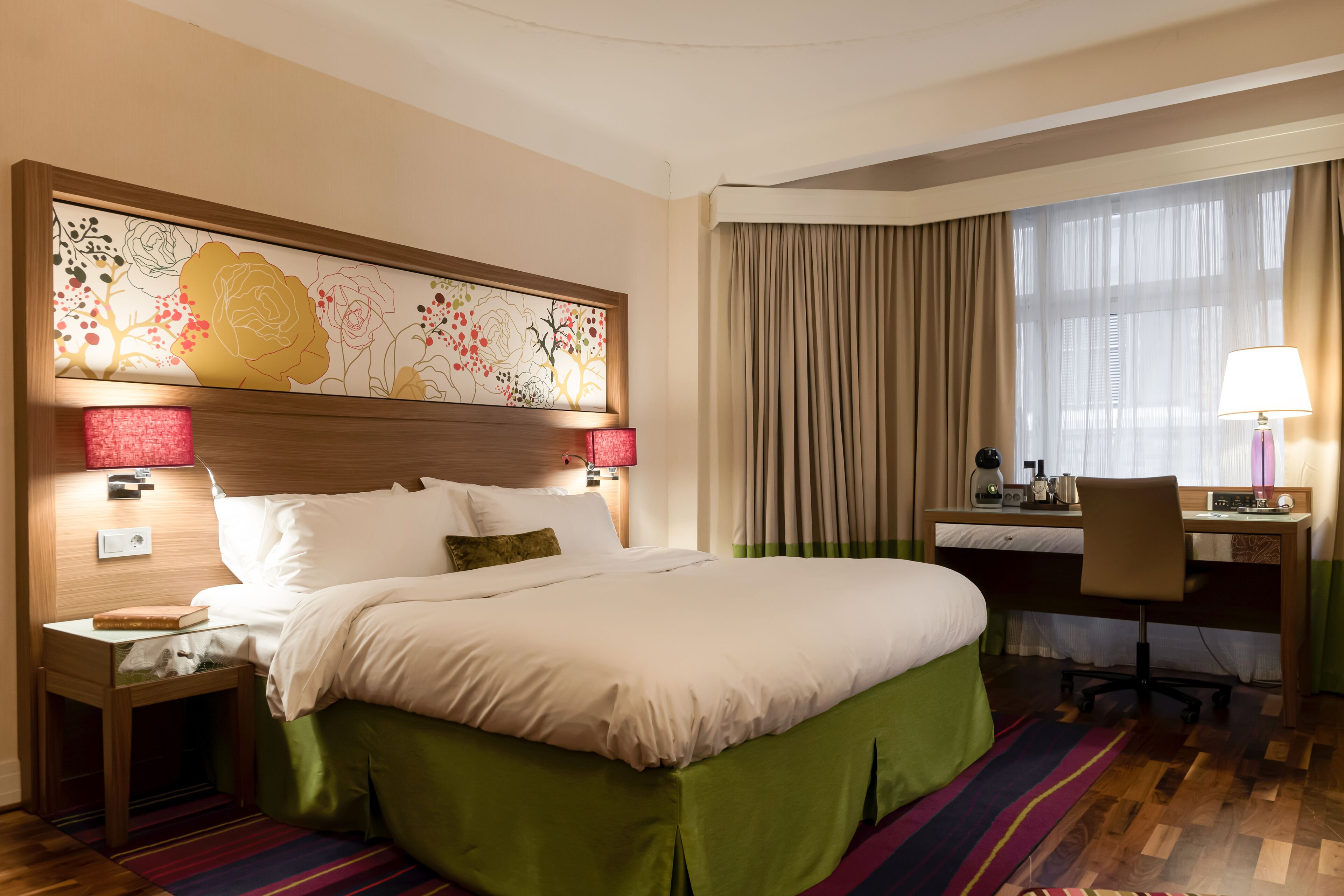 A hotel room with a large bed, a desk, a chair, a lamp, and a decorative wall piece.