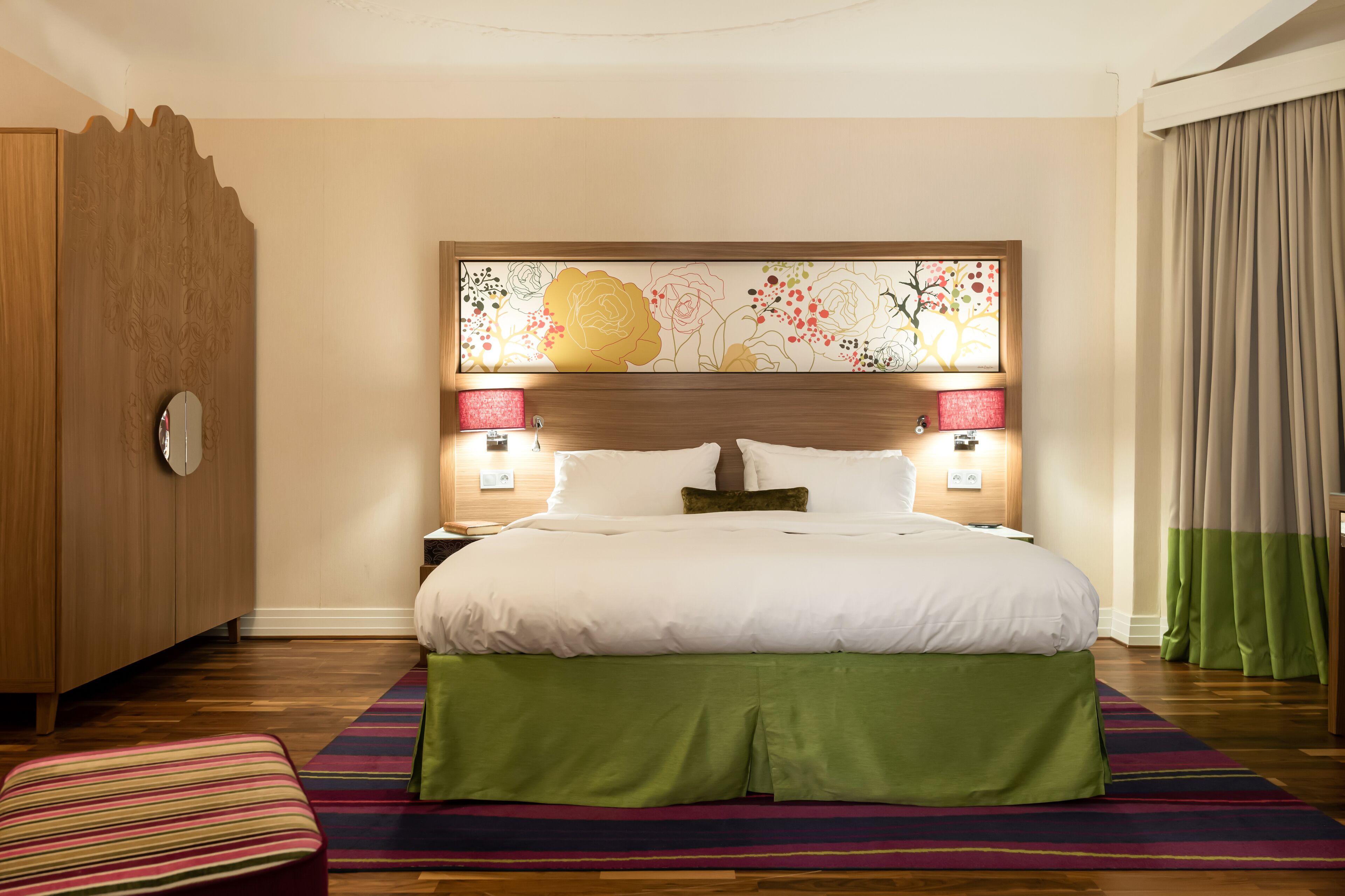 Quarto de hotel com uma cama grande, um guarda-roupa de madeira, uma mesa de cabeceira com abajur e um tapete listrado e colorido.