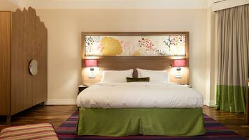 Quarto de hotel com uma cama grande, um guarda-roupa de madeira, uma mesa de cabeceira com abajur e um tapete listrado e colorido.