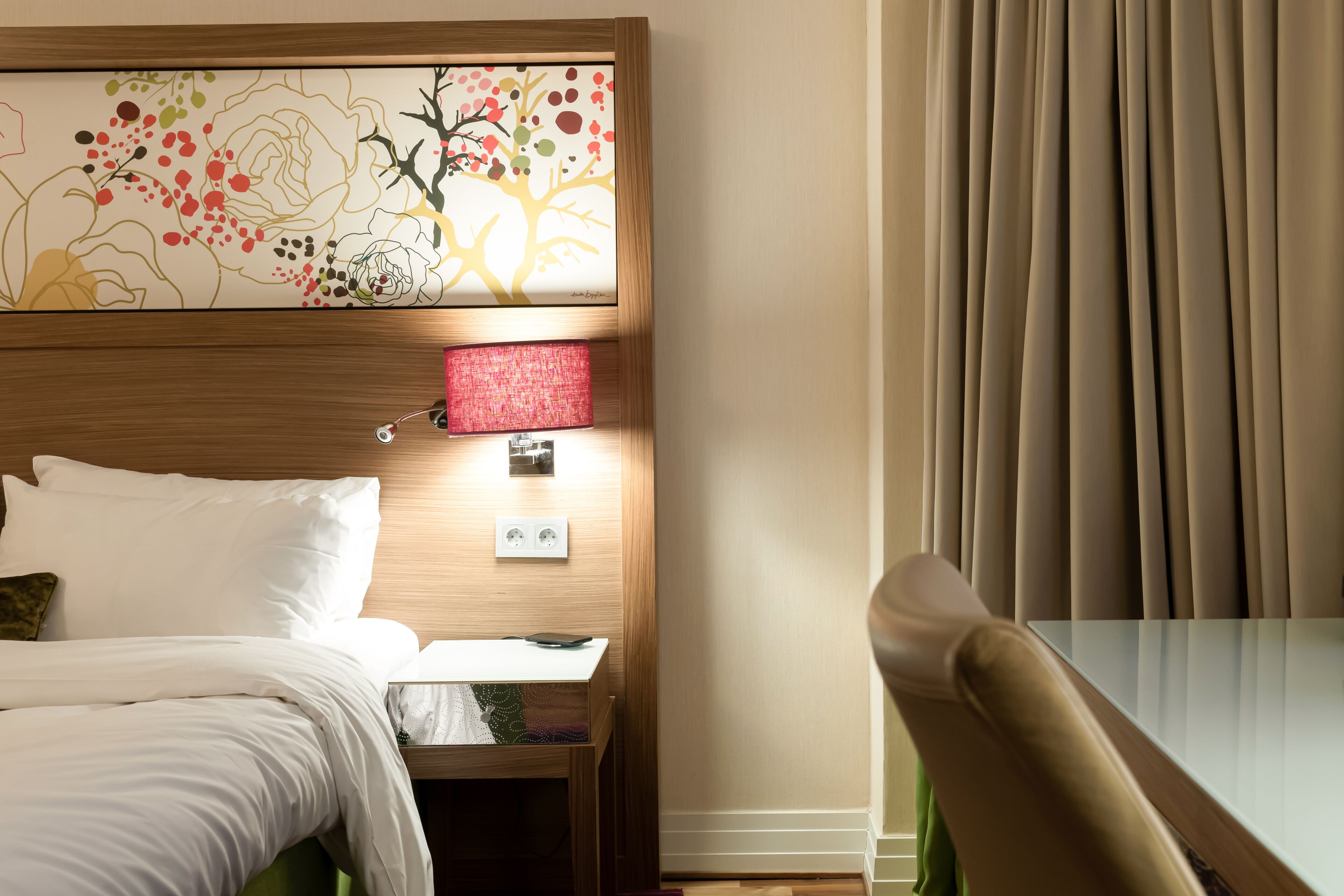 A hotel room with a bed, a nightstand, a table, a chair, a wall-mounted lamp, and a window with curtains.