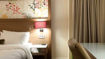 A hotel room with a bed, a nightstand, a table, a chair, a wall-mounted lamp, and a window with curtains.