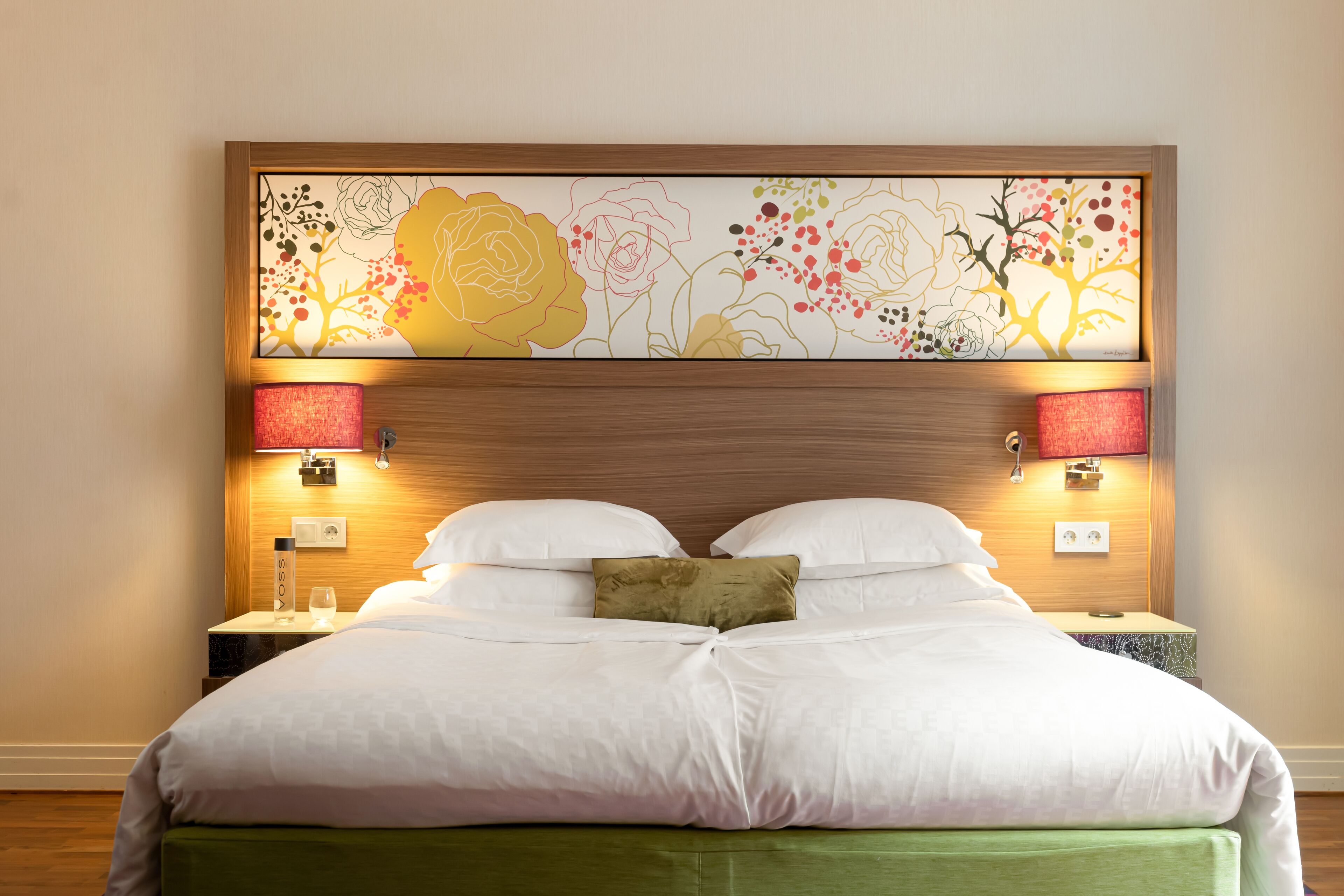 Una cama bien hecha con ropa blanca y detalles en verde, un cabecero de madera con una obra de arte floral, dos lámparas de noche y una mesita de noche con una botella y un vaso.