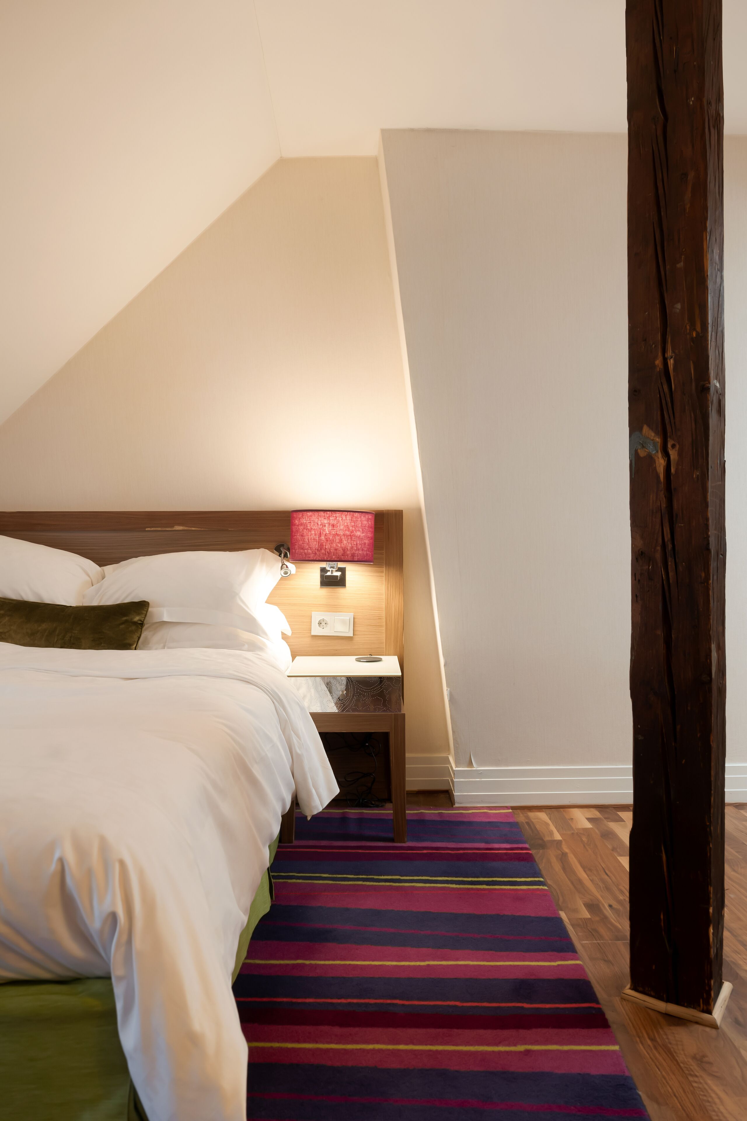 A bedroom with a bed, a nightstand, a lamp, and a colorful striped rug.