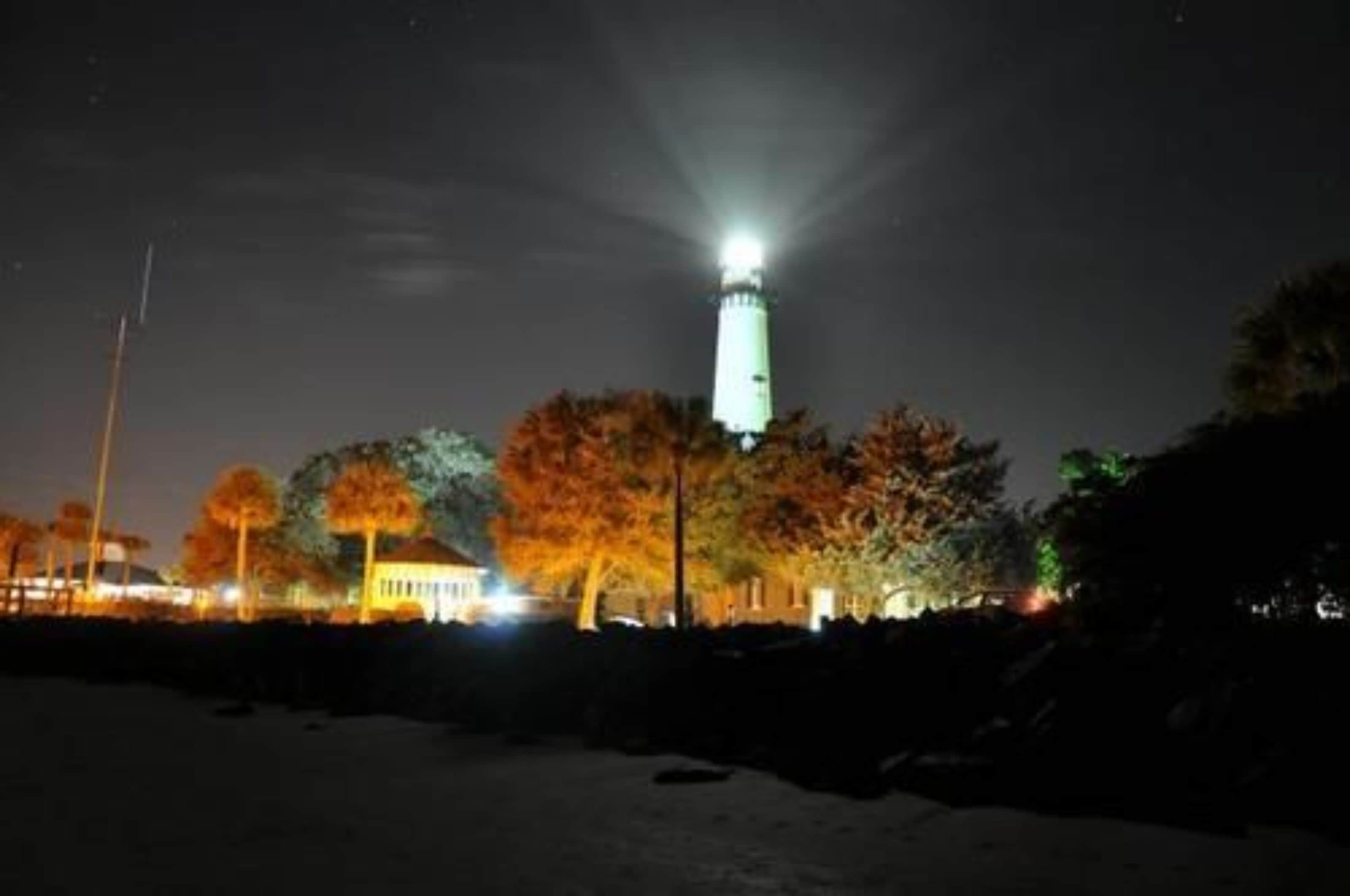 Un faro di notte con una luce intensa in cima, circondato da alberi con foglie autunnali.