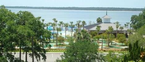 A view of a hotel with a water body in the background, surrounded by greenery and a parking area.