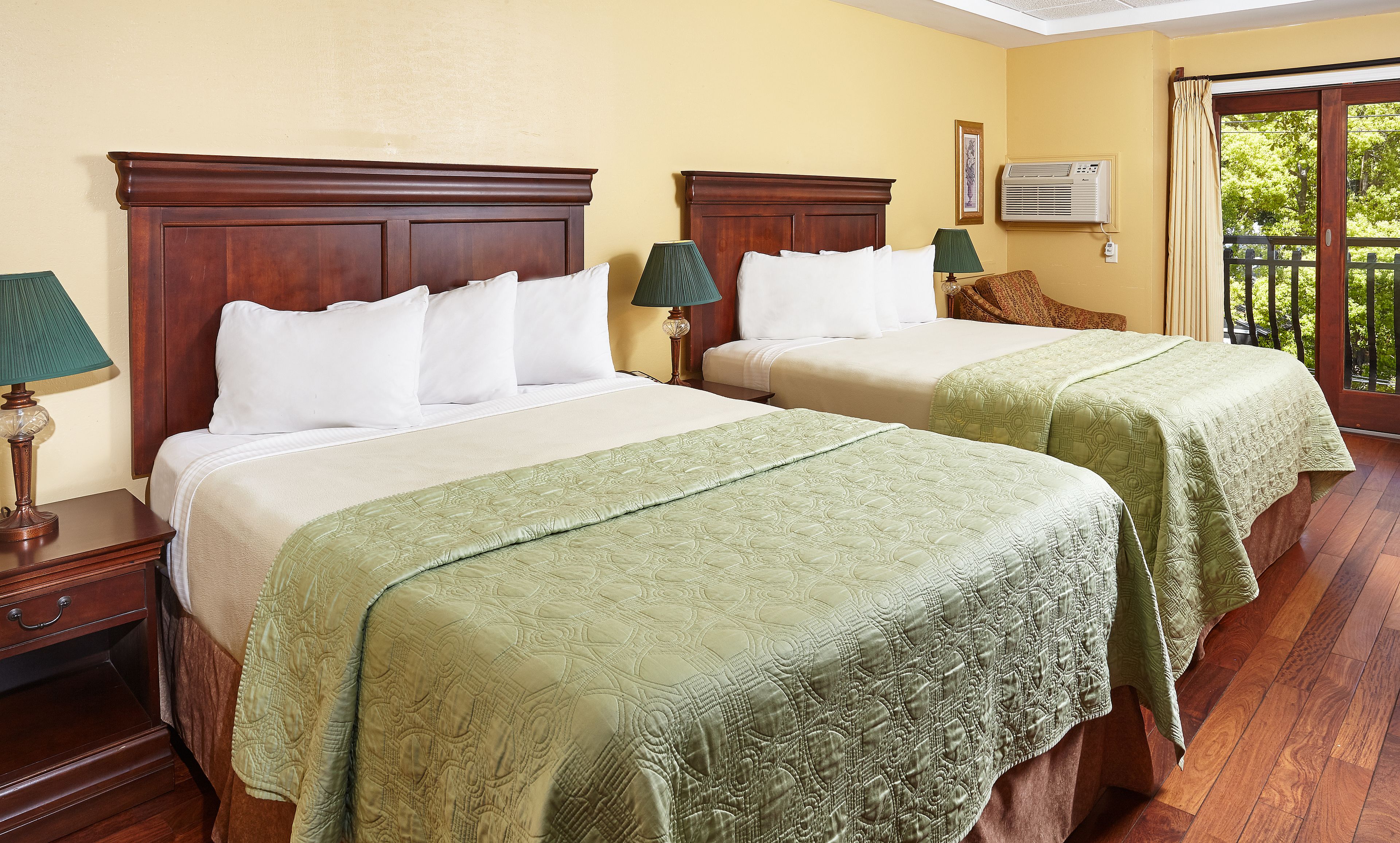 Une chambre d’hôtel avec deux lits, des têtes de lit en bois, des couvre-lits verts, une table de chevet avec une lampe, une petite table et une vue sur l’extérieur.