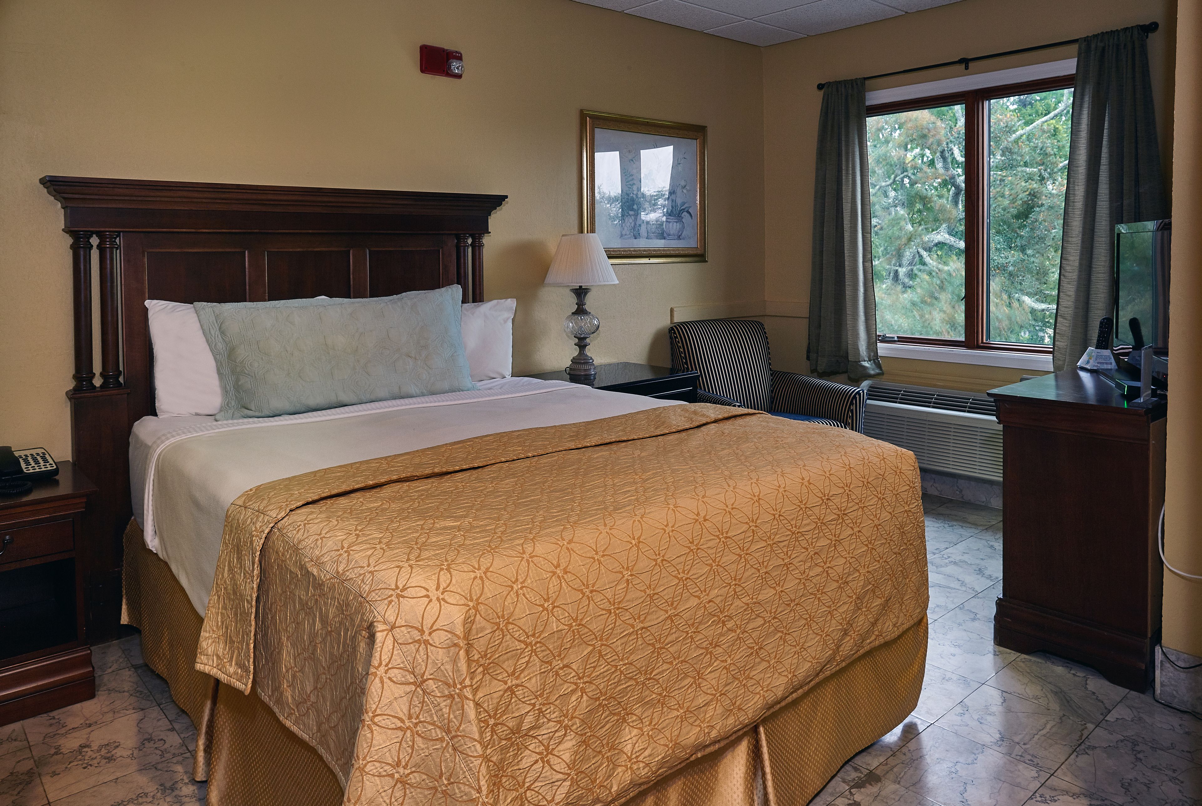 A hotel room with a large bed, a nightstand with a lamp, a desk with a computer, and a window with curtains.