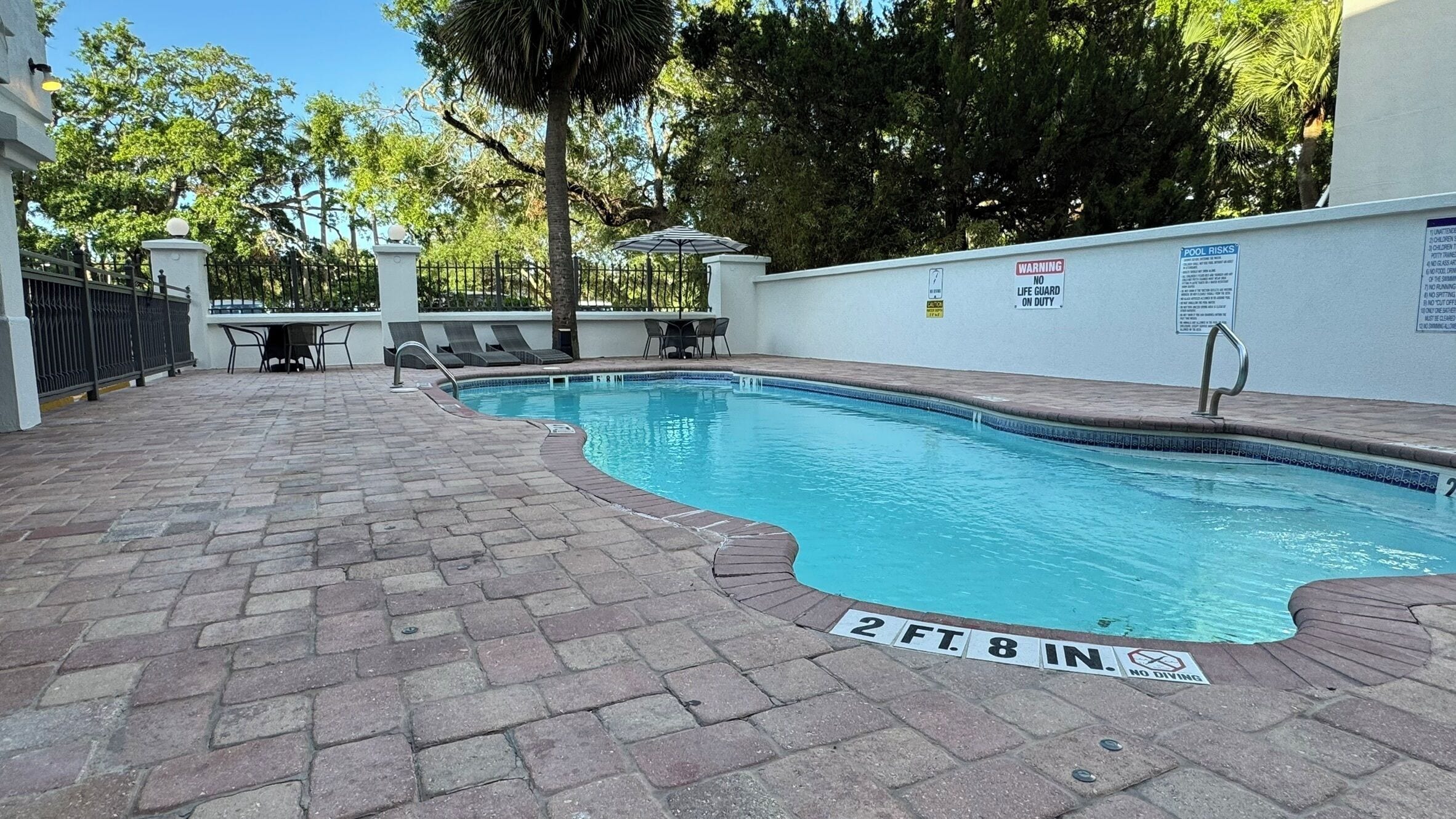 A rectangular pool with a brick deck, surrounded by a white wall and a metal fence. There are lounge chairs and a table near the pool.