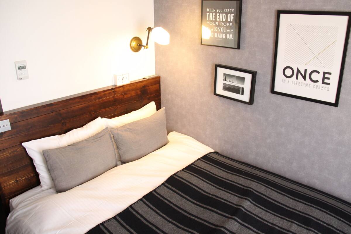 Una cama bien tendida con una manta a rayas, un cabecero de madera y una lámpara fijada a la pared. Hay cuadros enmarcados y una frase motivacional en la pared.