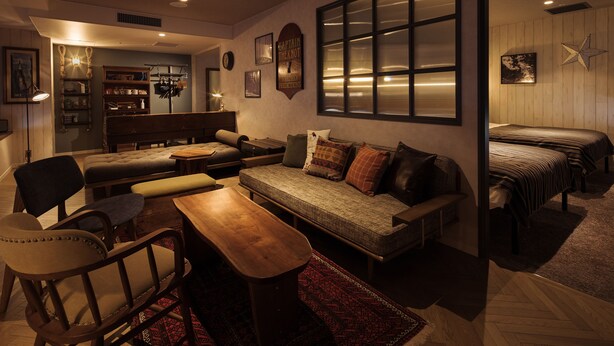 A cozy living room with a sofa, chairs, a wooden coffee table, and a bookshelf. There is a large window with blinds, a star decoration on the wall, and a clock. - Hotel The Knot Yokohama (Yokohama)