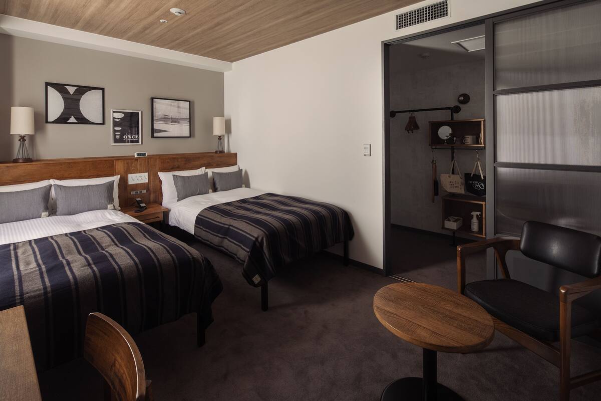 Habitación de hotel con dos camas, cabecera de madera, una mesita, una silla y una puerta corredera que da a un baño.