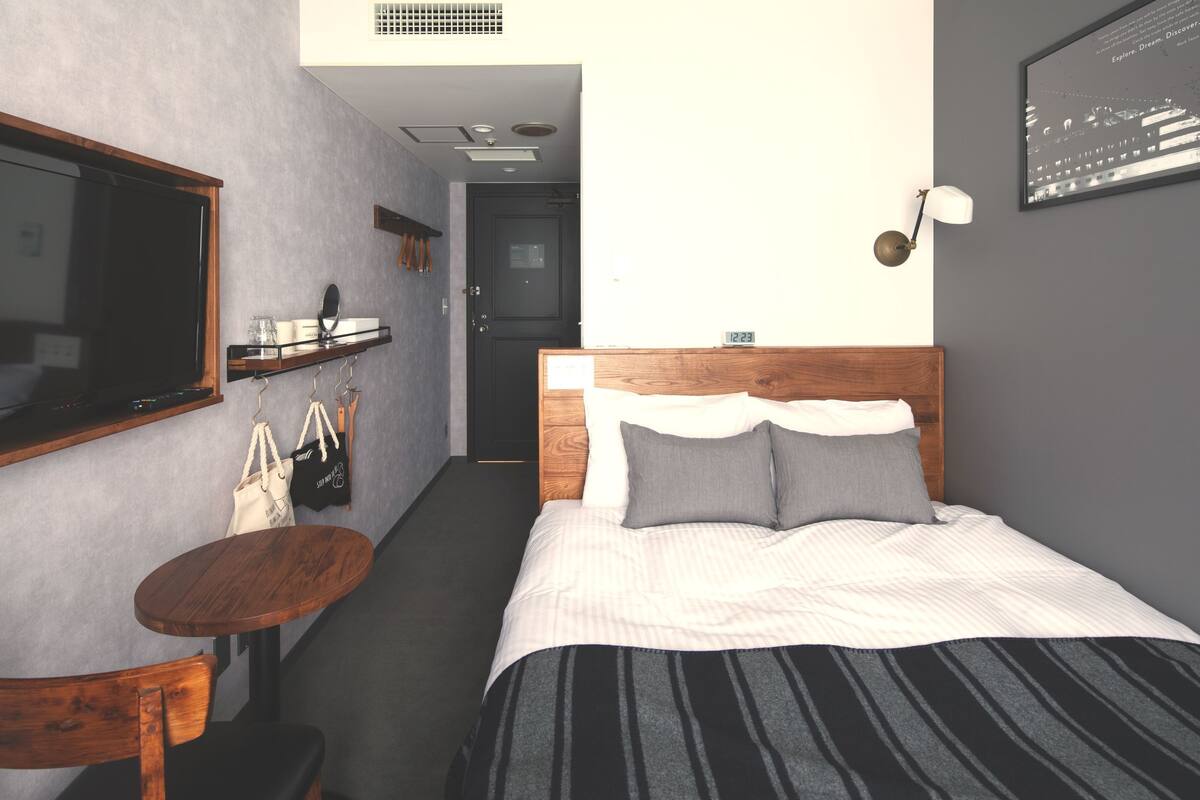 Habitación de hotel con una cama, una pequeña mesa de madera, una silla, un espejo, una estantería con maletas y una puerta que da a otra habitación.