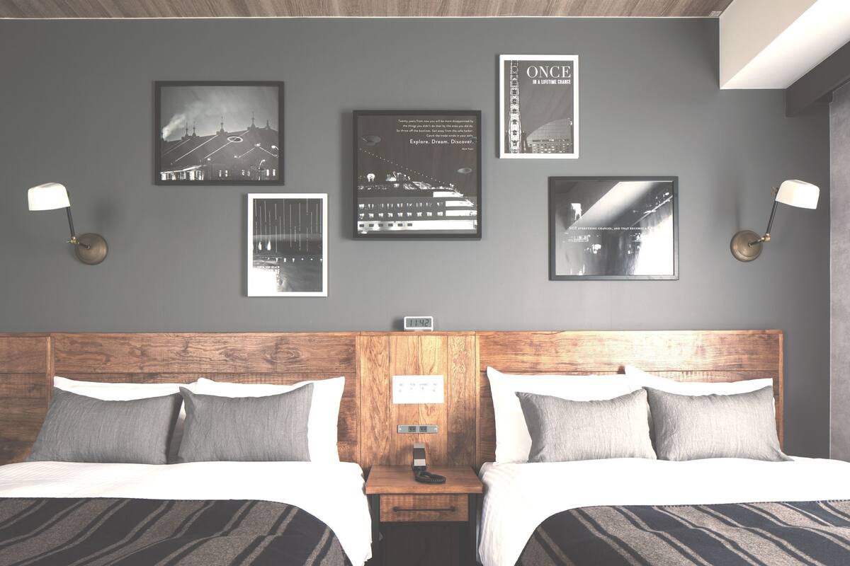 Habitación de hotel con cabecera de madera, dos camas, una mesita de noche y una obra de arte colgada en la pared.